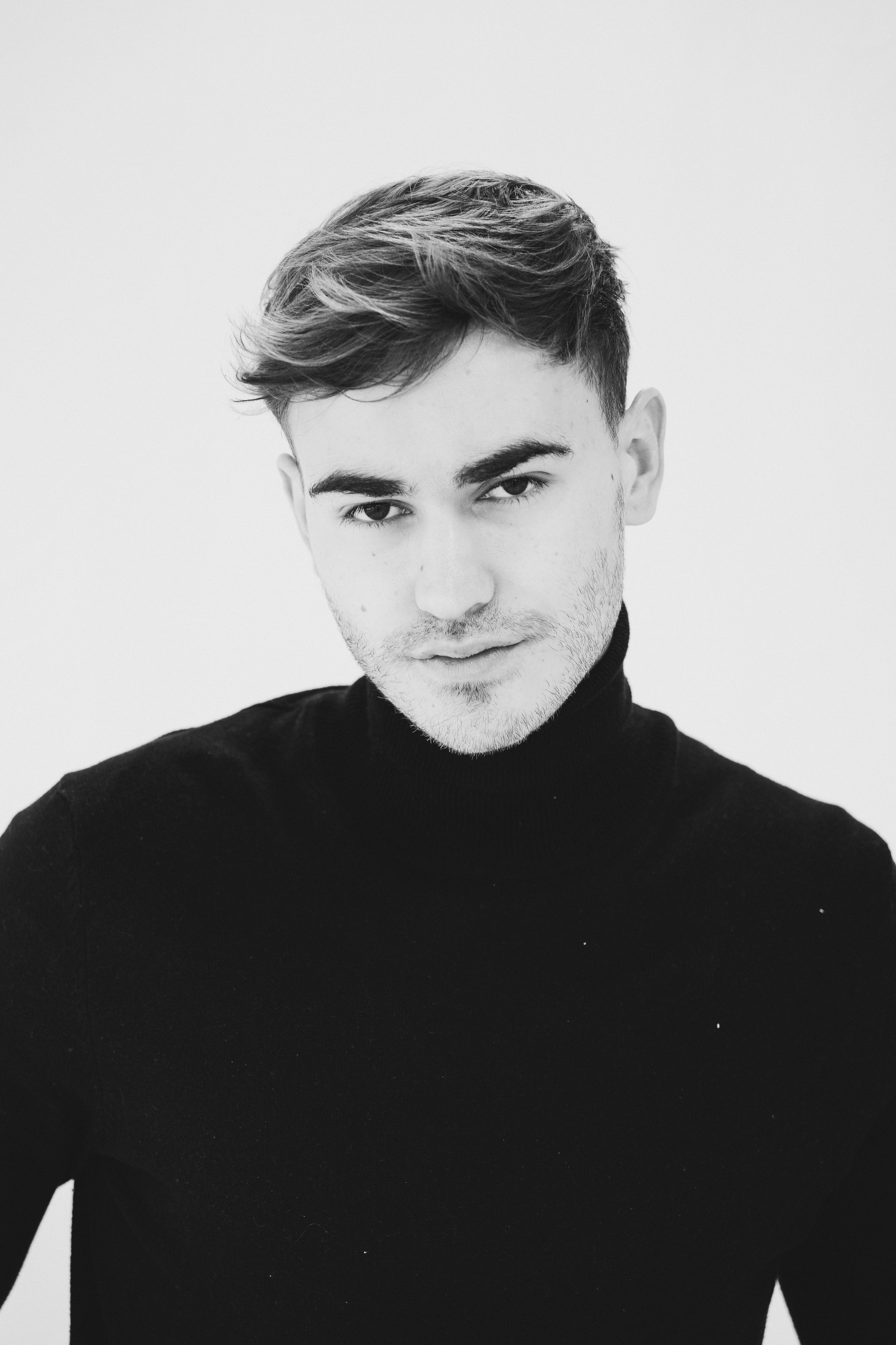 Black and white portrait of a young male model in a black turtleneck, looking confidently at the camera against a white background. Studio headshot of male model in black turtleneck, black and white fashion photography.