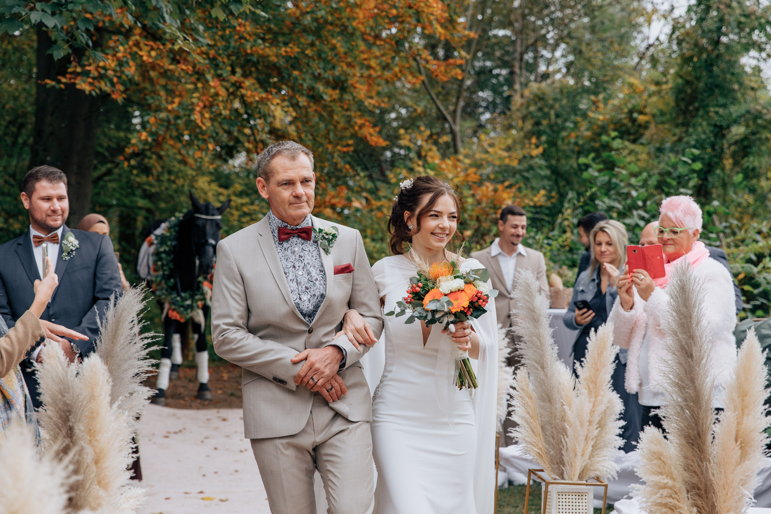 Der Vater führt die Braut zum Bräutigam, beide lächeln und sind nervös. Die anderen Gäste schauen ihnen zu.