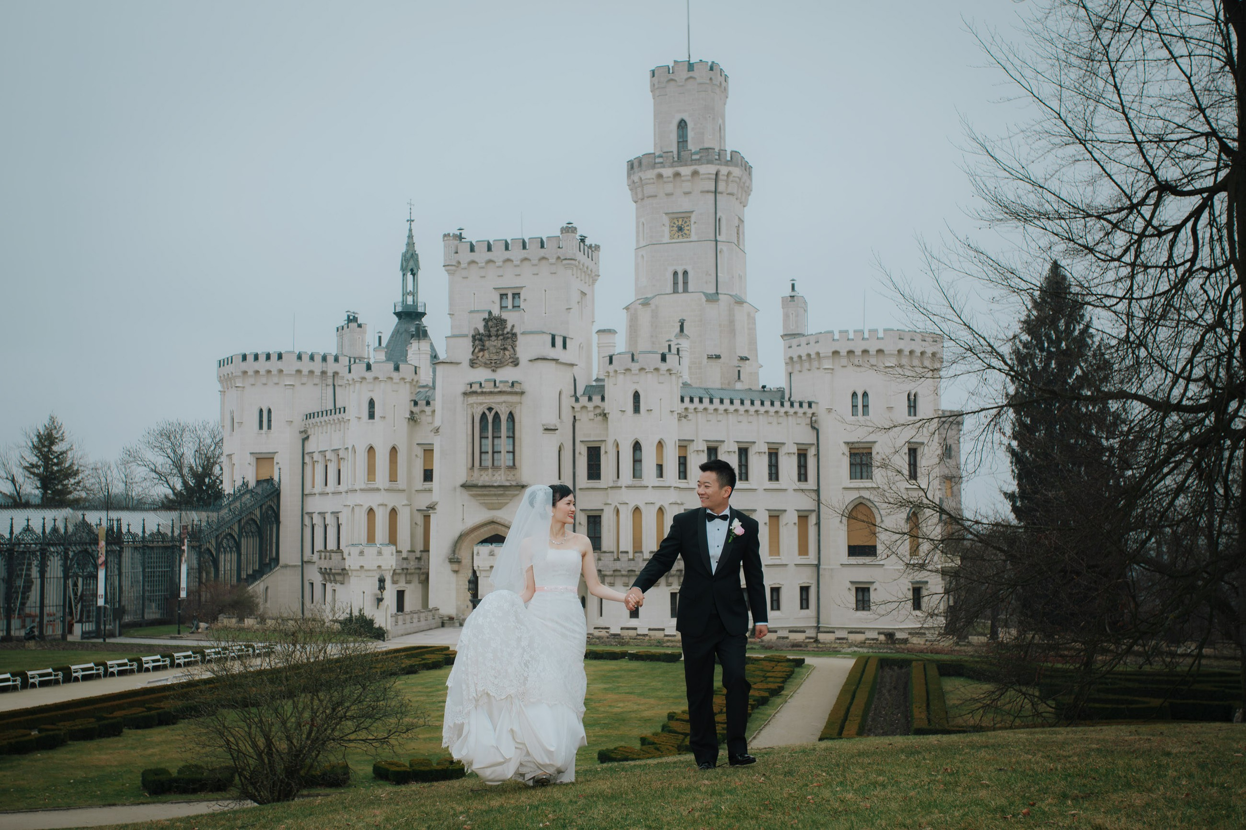 A groom wearing a black tuxedo takes his Hong Kong bride by her hand as they walk around the grounds of the historic Castle Hluboka where they had earlier held their destination wedding.