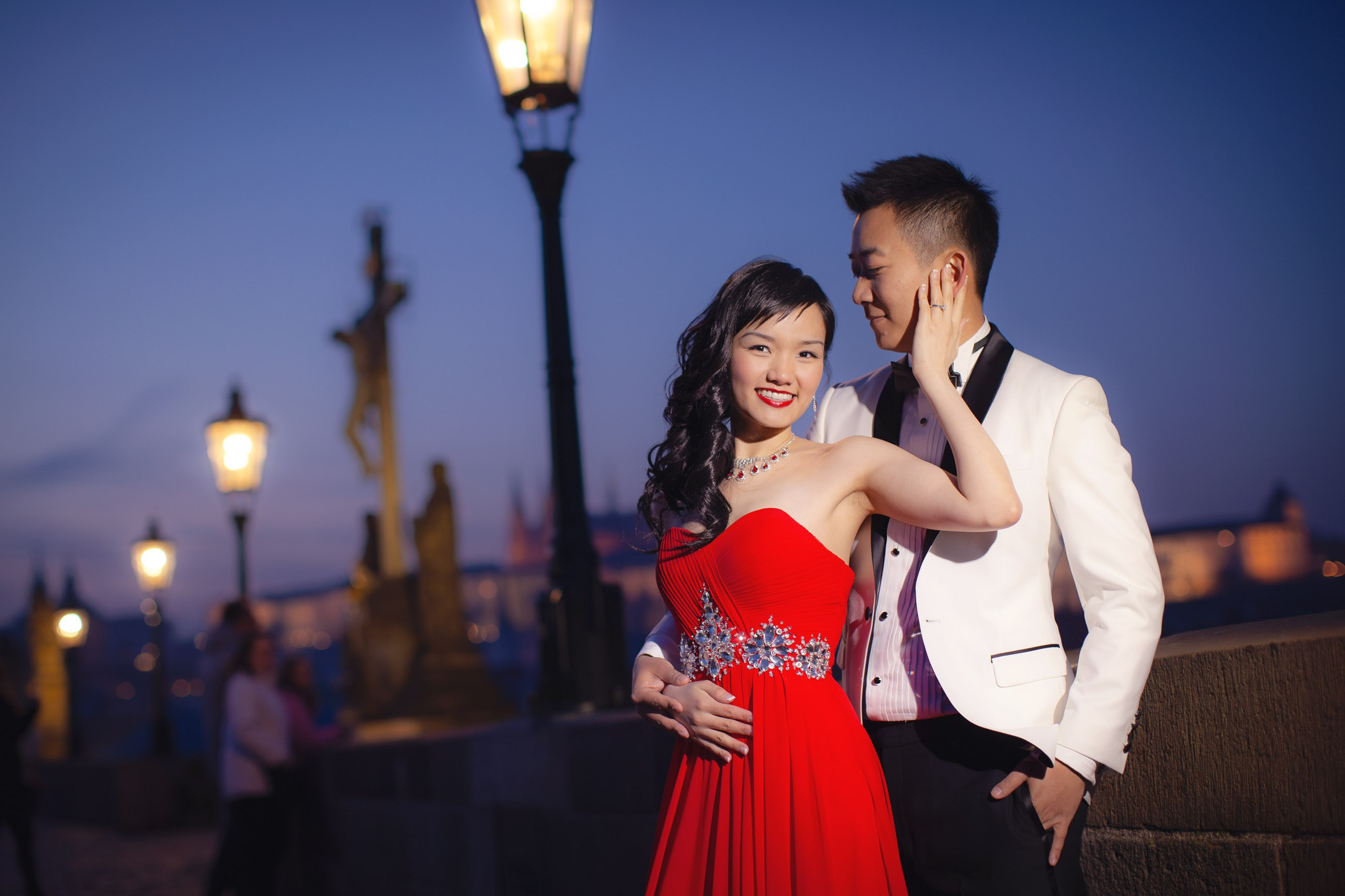A white tuxedo wearing man is caressed by a woman wearing an elegant red evening dress underneath the historic gas lamps along the Charles Bridge at night.