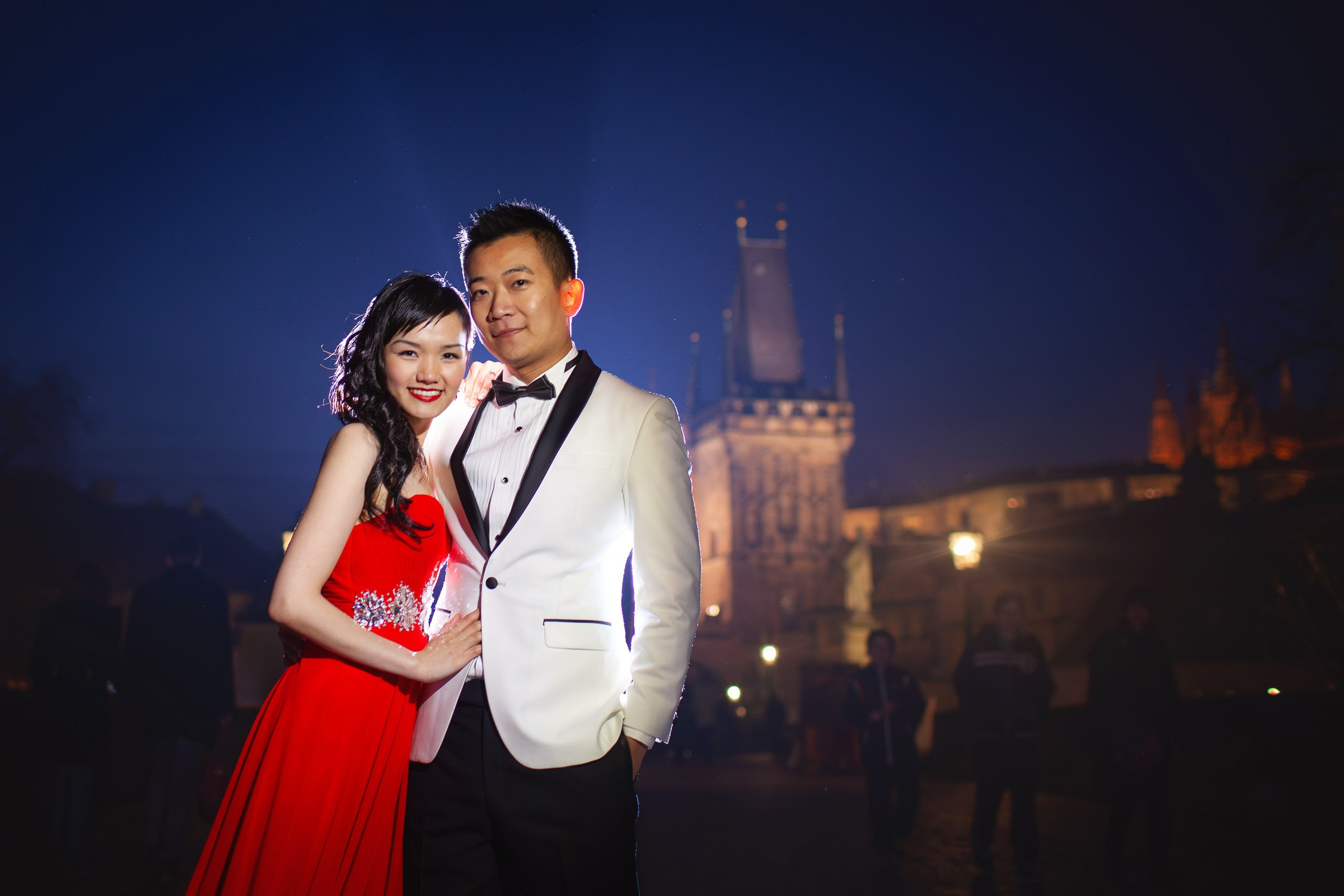 A smiling Hong Kong woman wearing a stylish red evening dress and her white tuxedo wearing man pose for a photo atop a misty Charles Bridge at night.