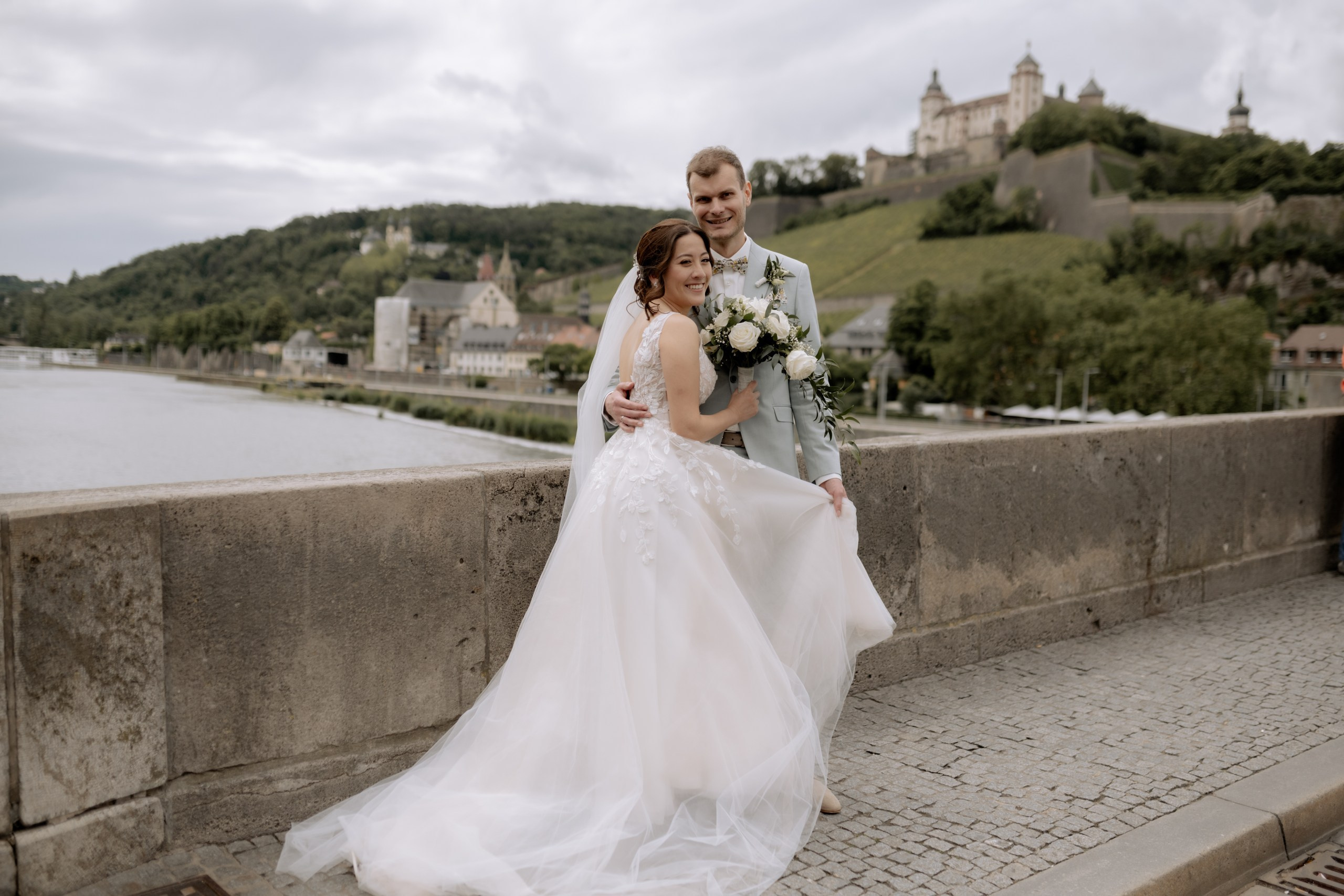 Die Braut lehnt sich an die Steinbrüstung der Alten Mainbrücke, schaut lachend zum Bräutigam. Im Hintergrund fließt ruhig der Main