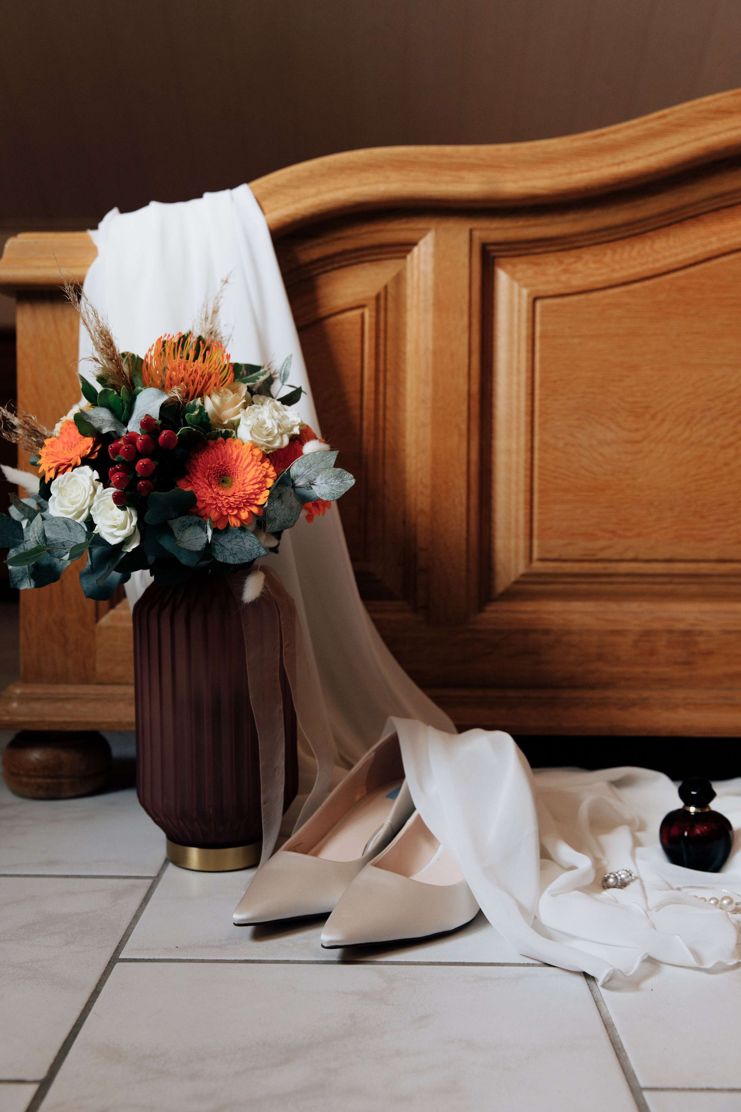 Herbstlicher Brautstrauß in warmen Farben in einer eleganten Vase, daneben Hochzeitsschuhe mit zartem Schleier, arrangiert vor einer rustikalen Holzwand für eine romantische Hochzeit.