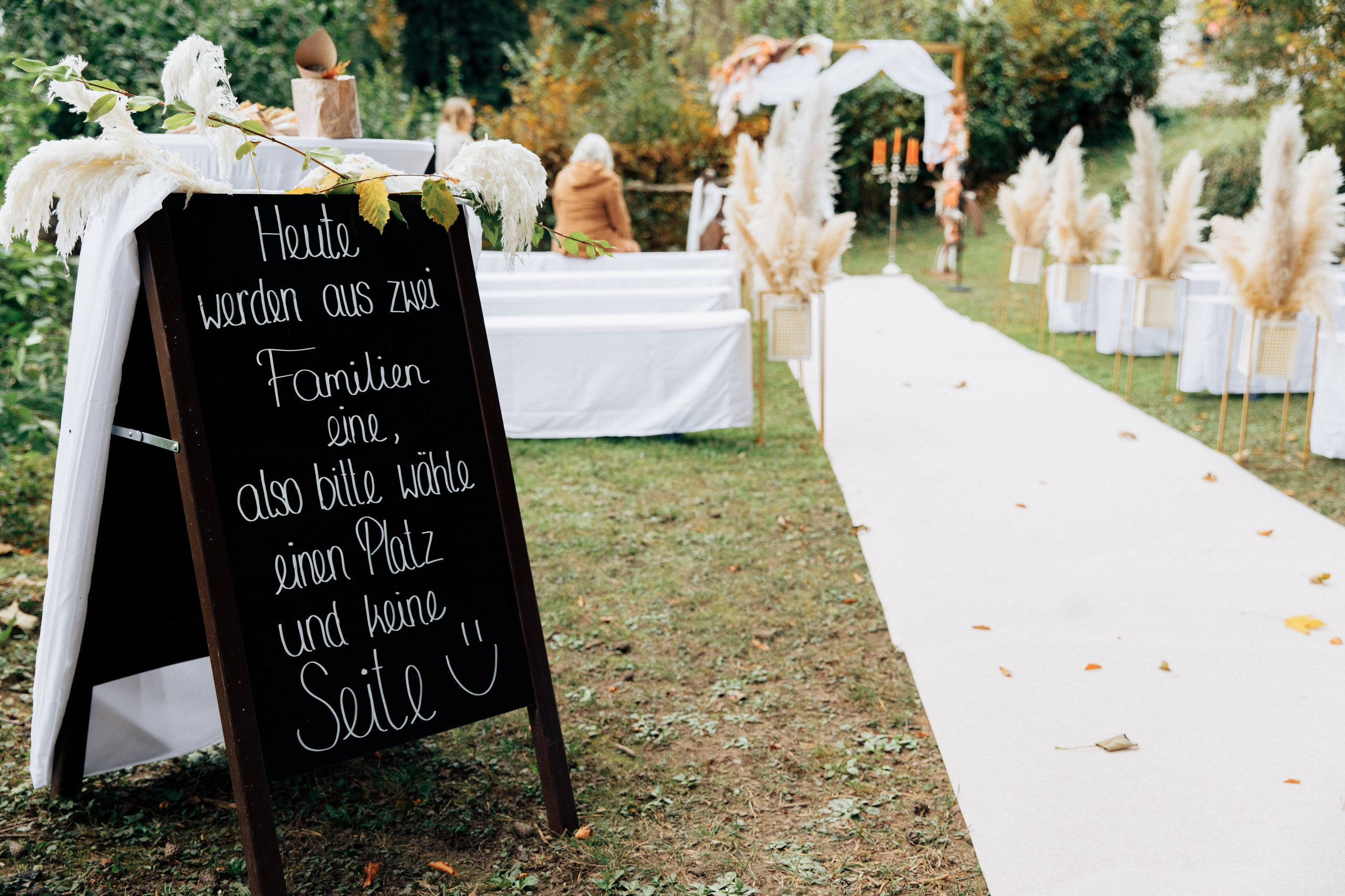 Im Vordergrund ein Schild mit einer niedlichen Aufschrift, im Hintergrund die Location, wo die Hochzeitszeremonie stattfinden wird, alles in herbstlichen Farben.