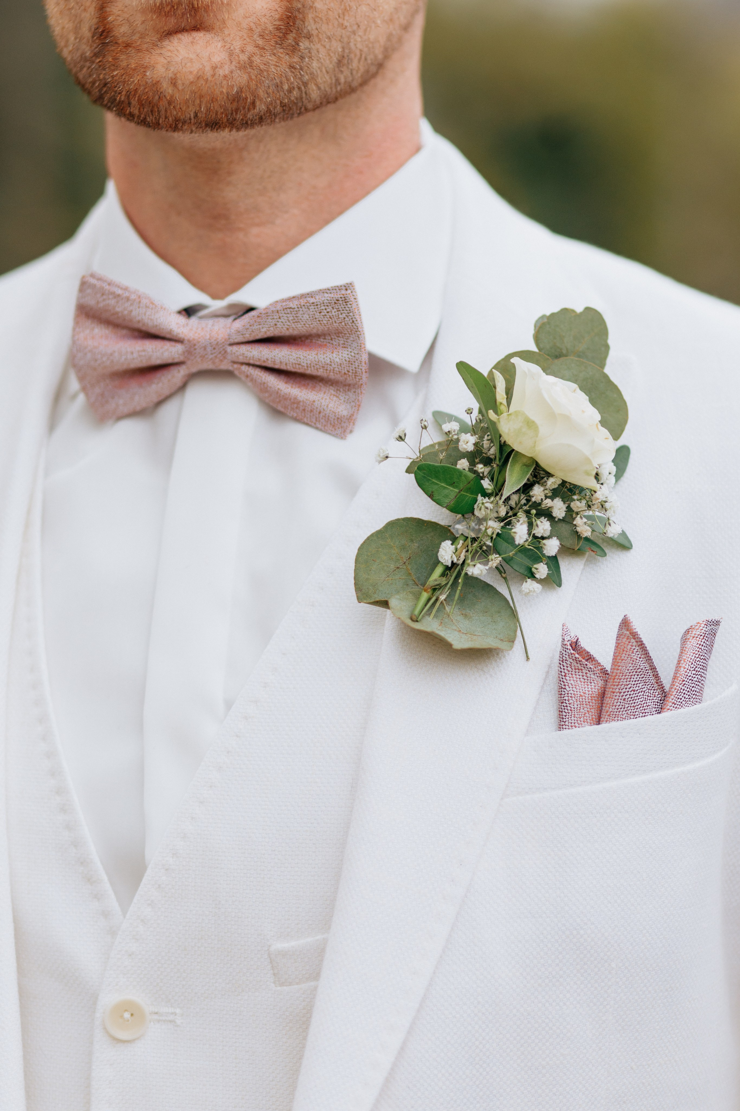 Elegantes Outfit des Bräutigams: Weißer Anzug, rosa Fliege und eine Ansteckblume mit Eukalyptus.