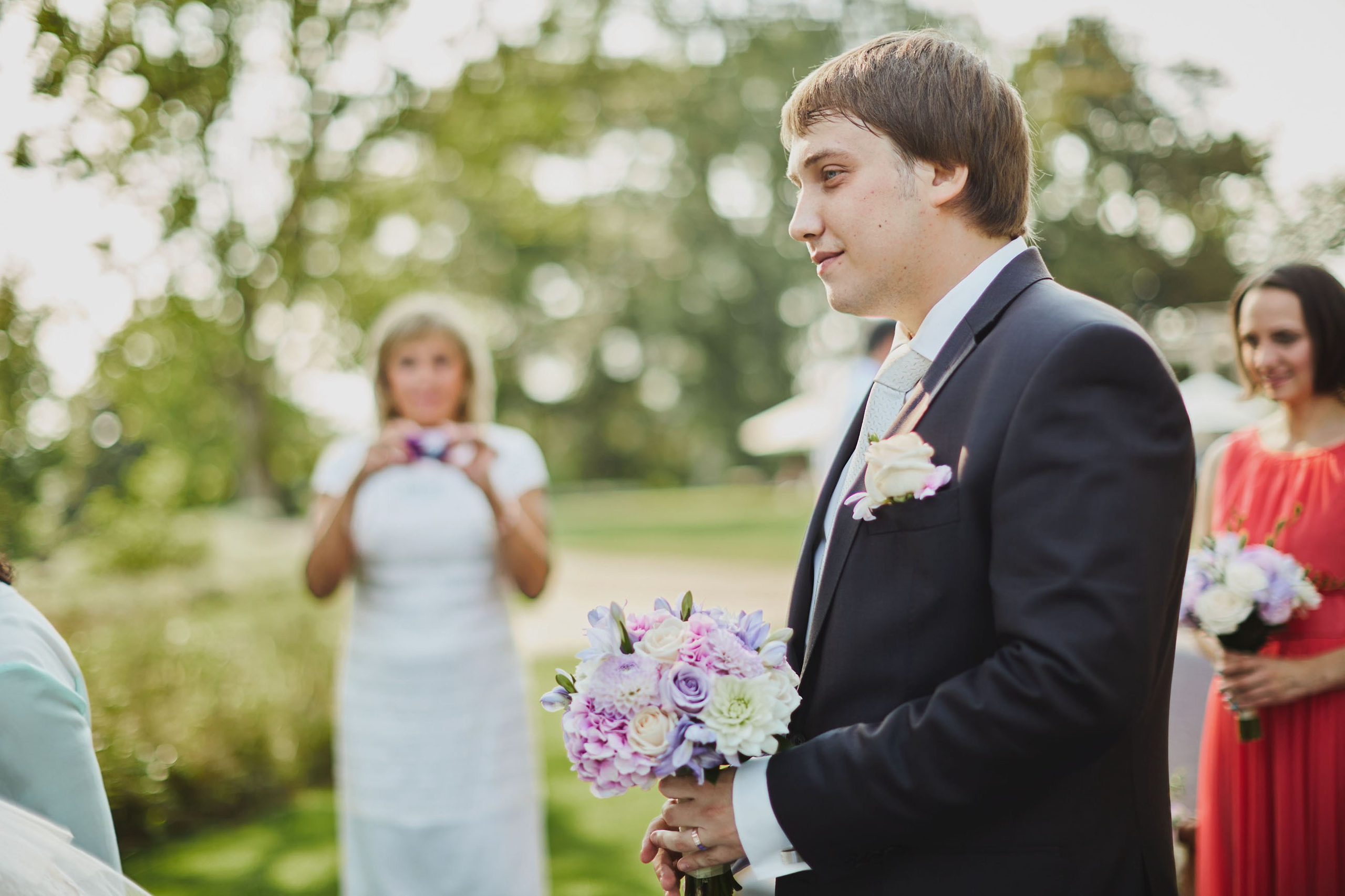 Groom with bouquet waiting as bride embraced by family at chateau wedding.