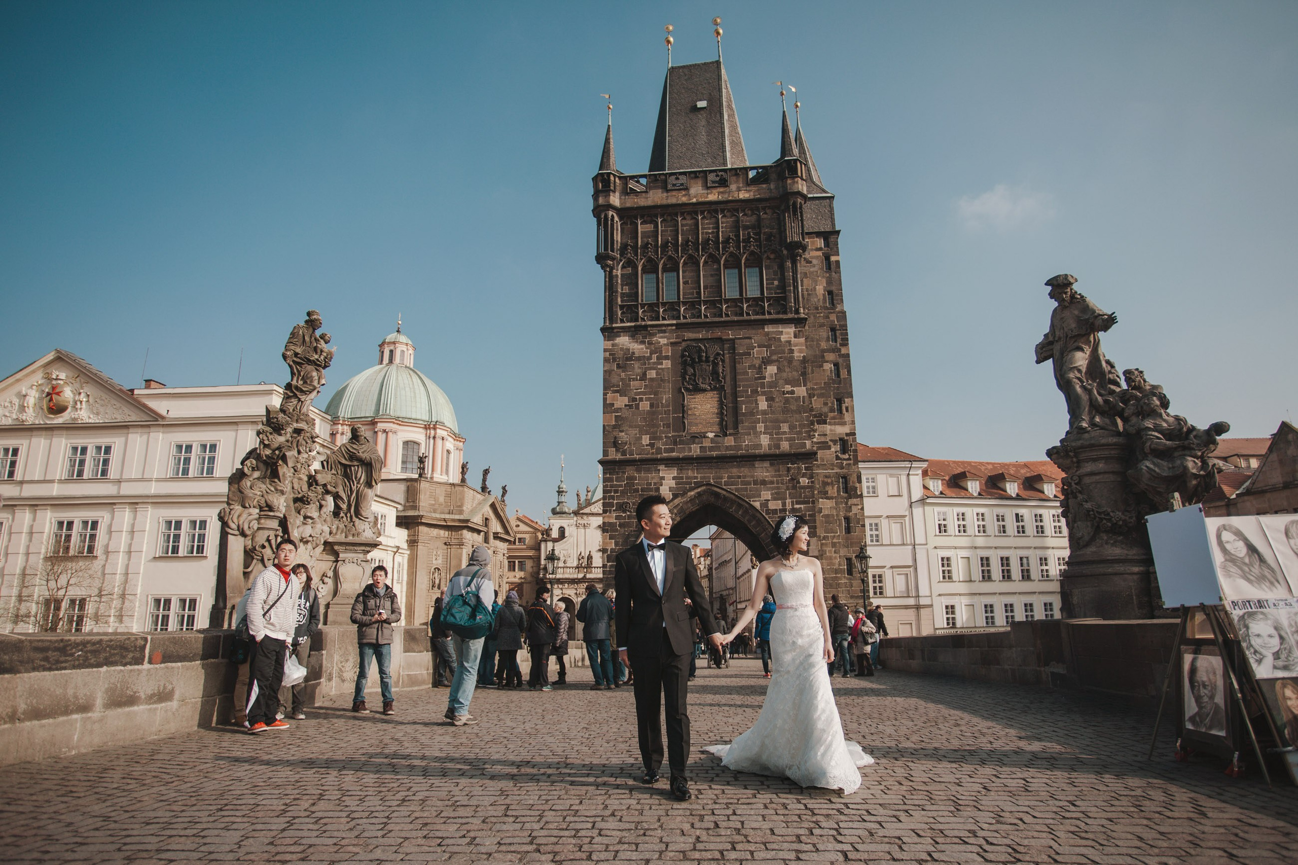 An elegant couple from Hong Kong dressed in stylish wedding attire walks hand in hand across the Charles Bridge during their winter love story session in Prague.