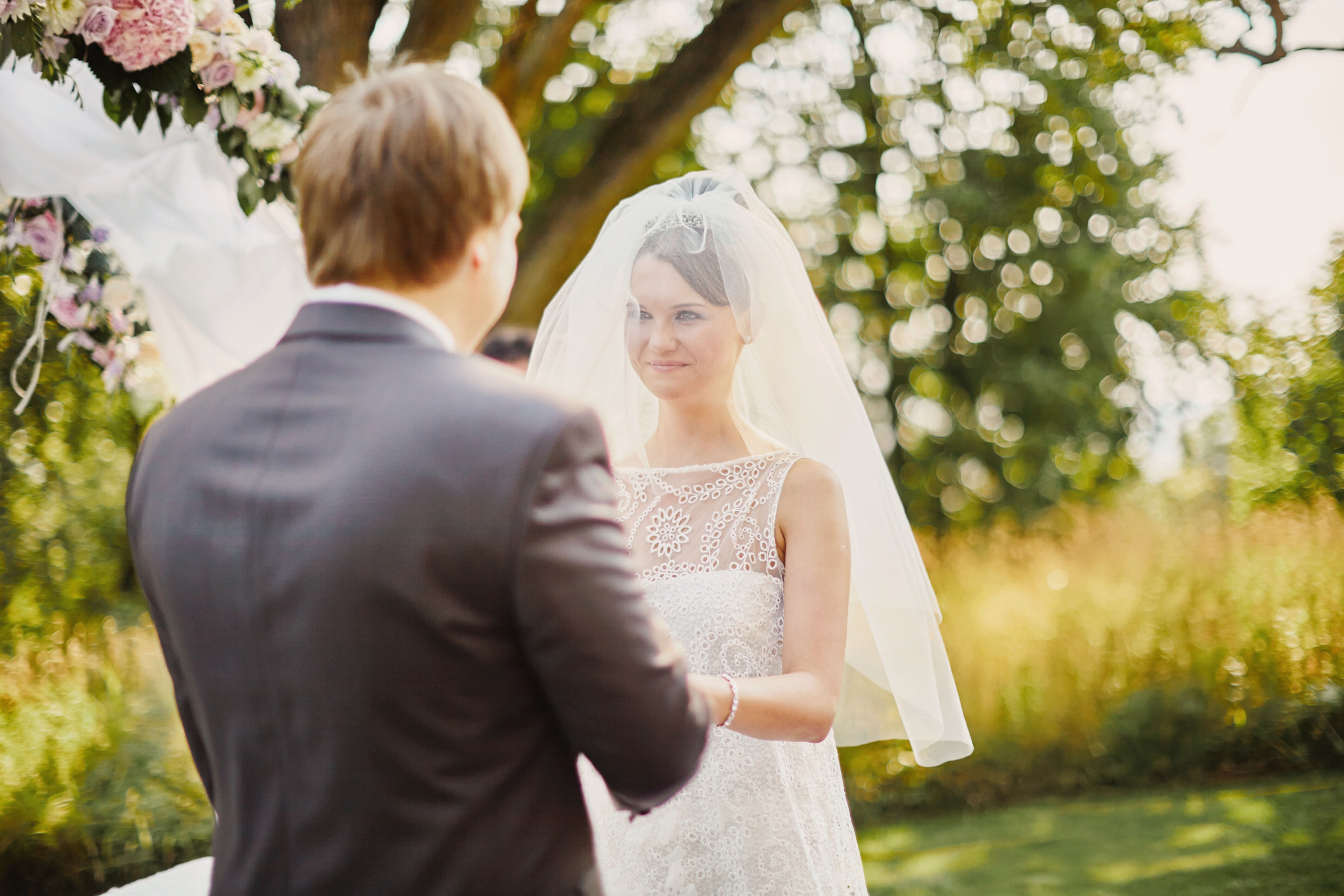 Bride holds hands with the groom as they stand near a floral arch during their summer, outdoor wedding on the grounds of a chateau in Czechia.