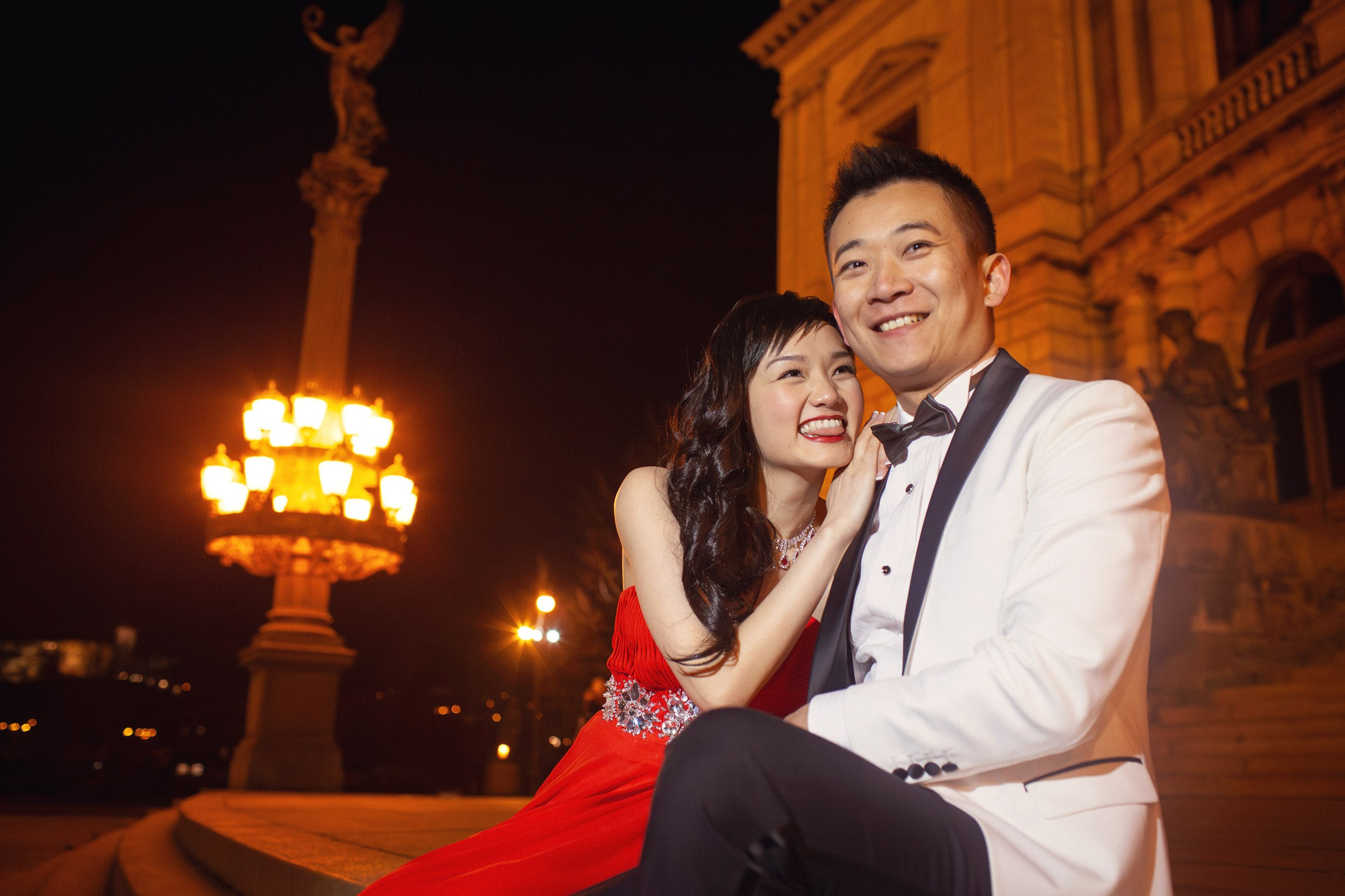 A radiant woman wearing a red evening dress leans into the lap of a man wearing a white tuxedo as they enjoy the sites and sounds of Prague from their vantage point at the Rudolfinum at night.