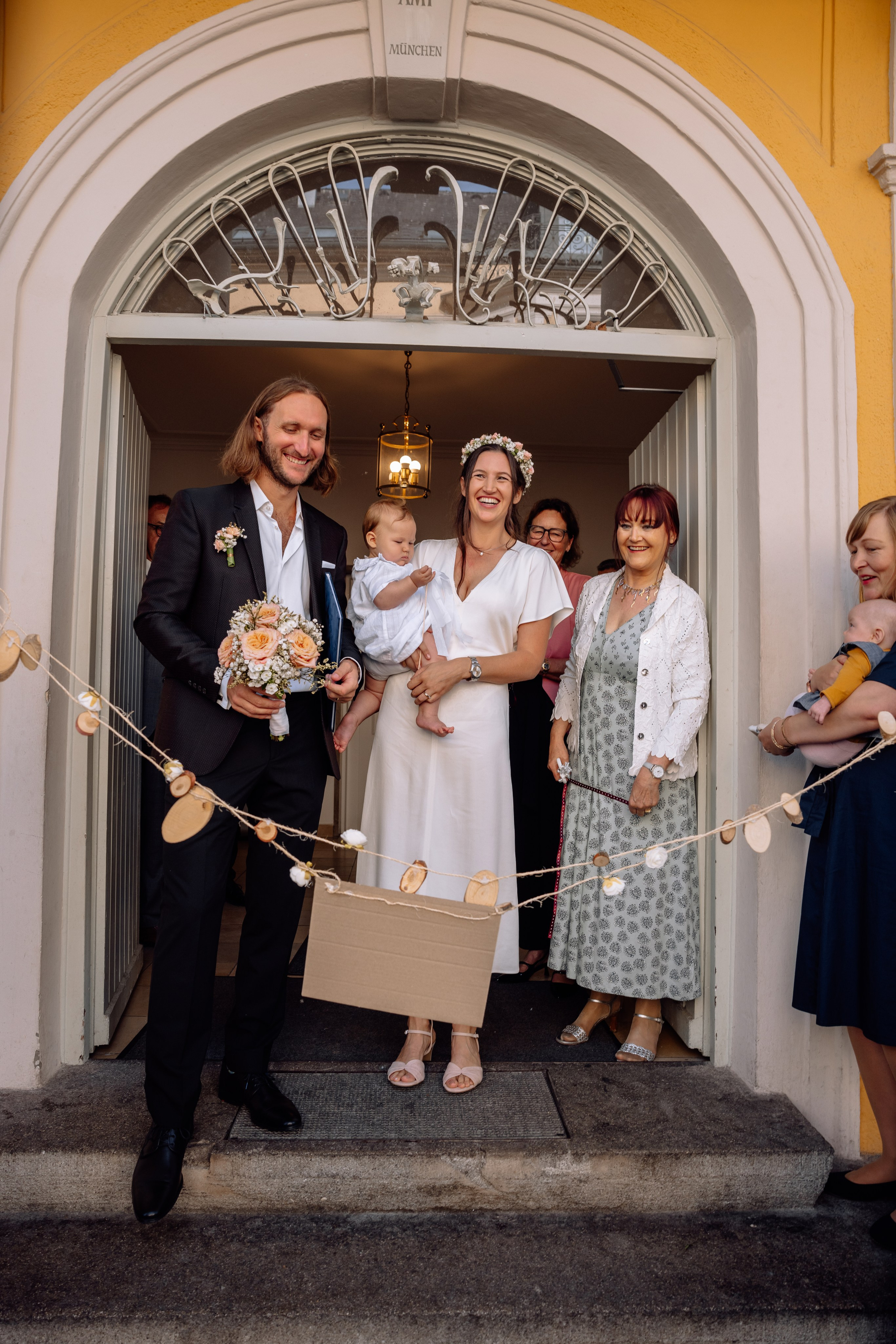 Braut und Bräutigam mit Gästen vor Standesamt Mandlstraße 14 – Baby in den Armen