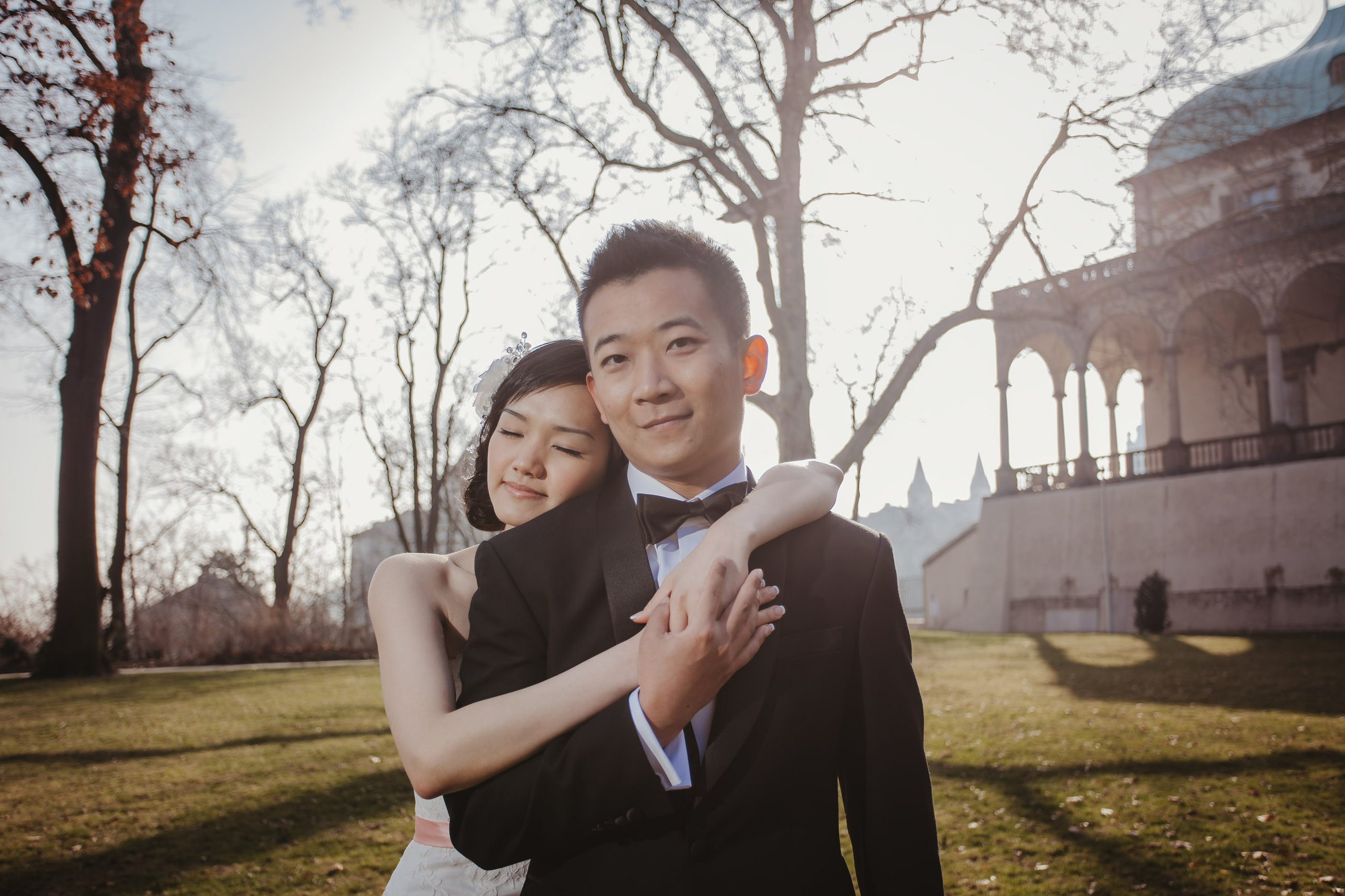 A stylish Hong Kong bride embraces her groom from behind as they stand near the Belvedere in Prague.