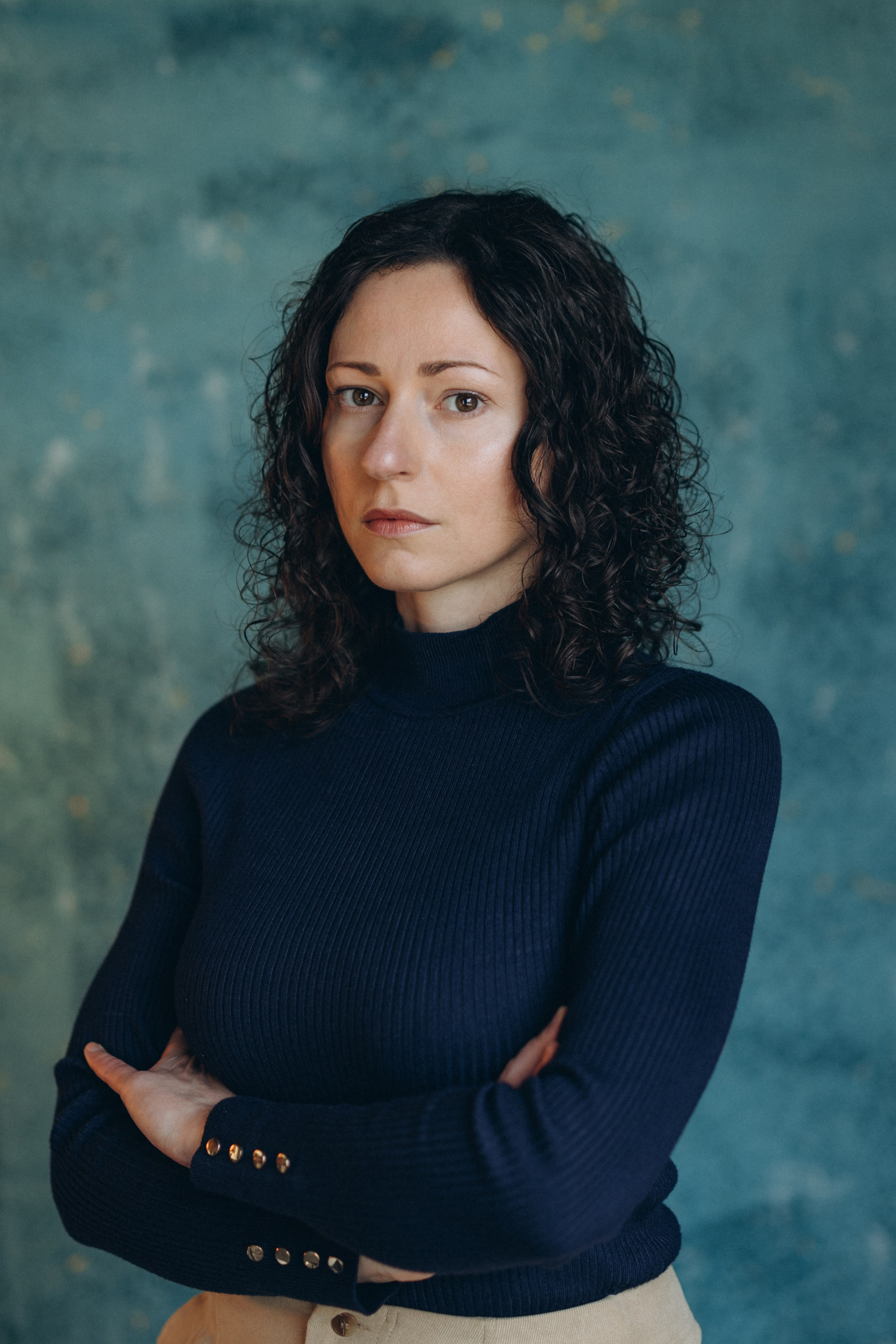 Actor headshot of a woman with curly hair in a navy blue top, arms crossed, looking down thoughtfully