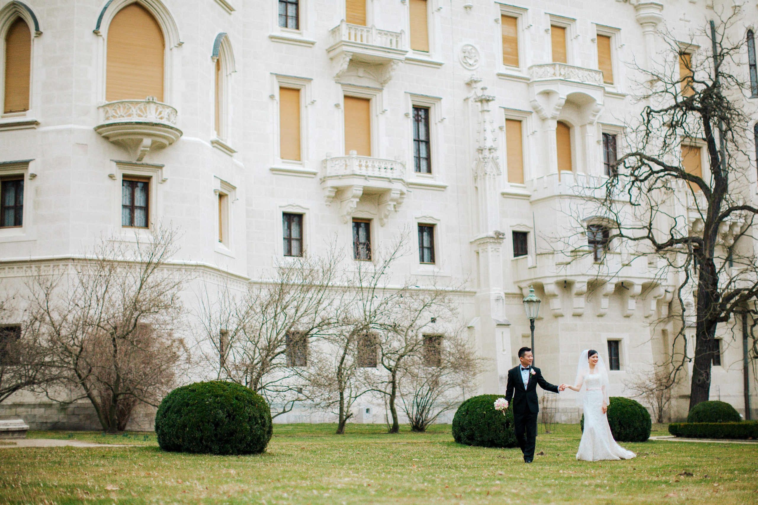 A smling Hong Kong bride & groom walk hand in hands on the historic grounds of the State Chateau of Hluboka where they had earlier held their intimate destination wedding.