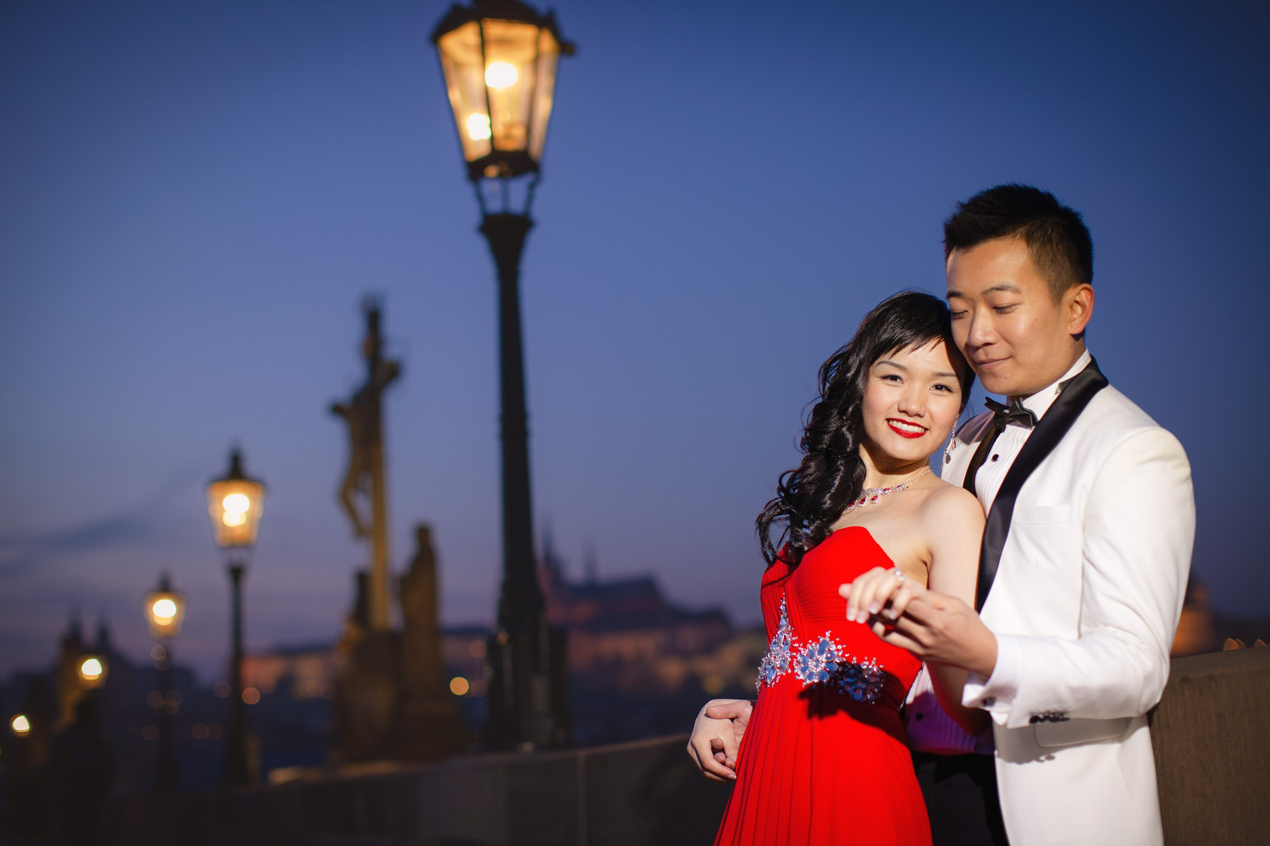 A woman in red wearing a stylish evening dress has her hand held by a white tuxedo wearing man during their night time portrait session atop the Charles Bridge.