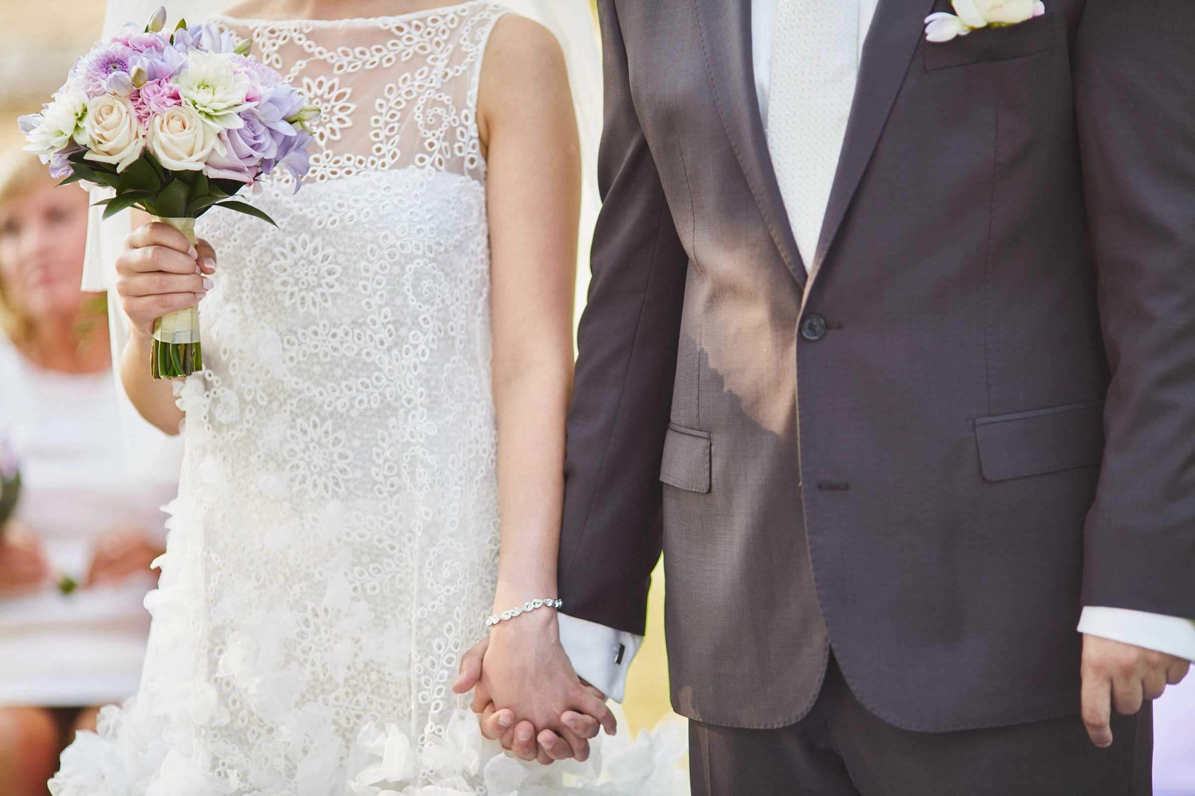 The brides and grooms hands as she holds her bouquet during their summer, outdoor wedding on the grounds of a chateau.