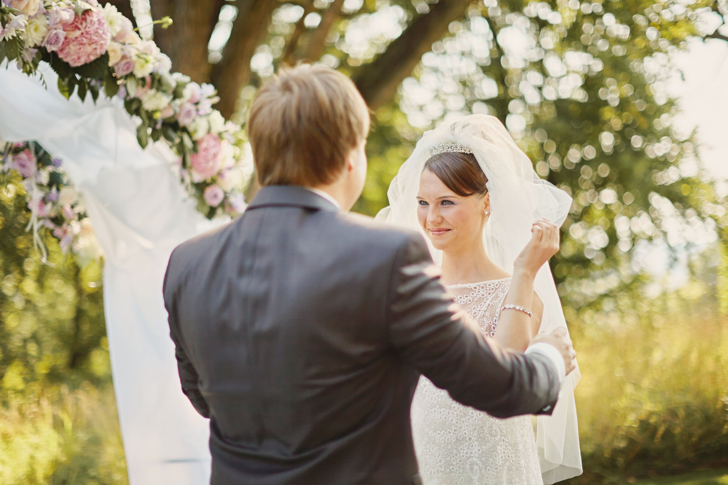 Bride is assisted by the groom as she lifts her veil under a floral arch during their summer, outdoor wedding on the grounds of a chateau in Czechia.