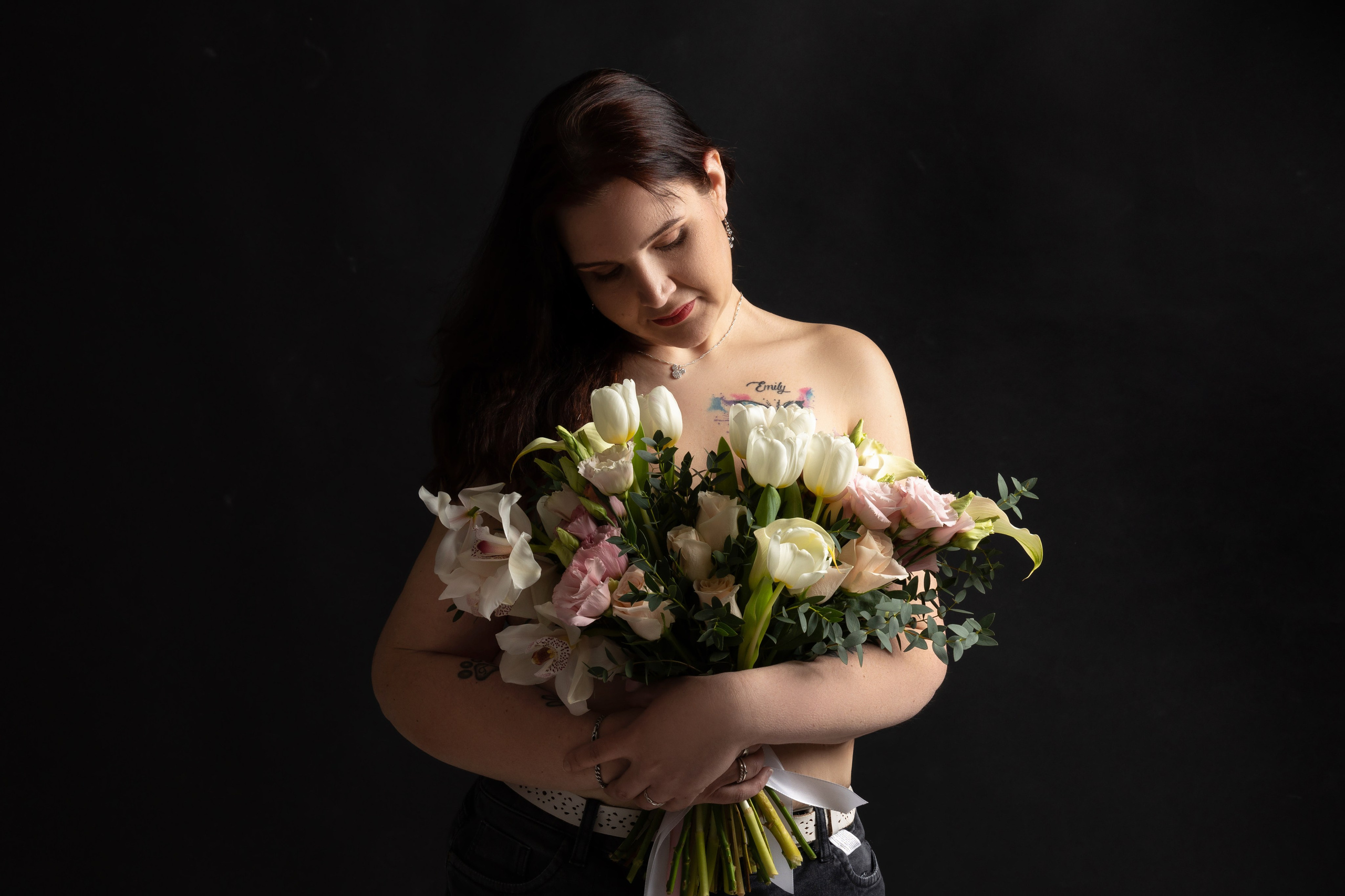 "Frau mit dunklen Haaren hält einen großen Strauß aus weißen und rosa Blumen im Arm. Sie blickt sanft nach unten, ihr Oberkörper ist teilweise entblößt. Das weiche Studiolicht hebt ihre zarte Haut und das florale Arrangement vor dem dunklen Hintergrund hervor."