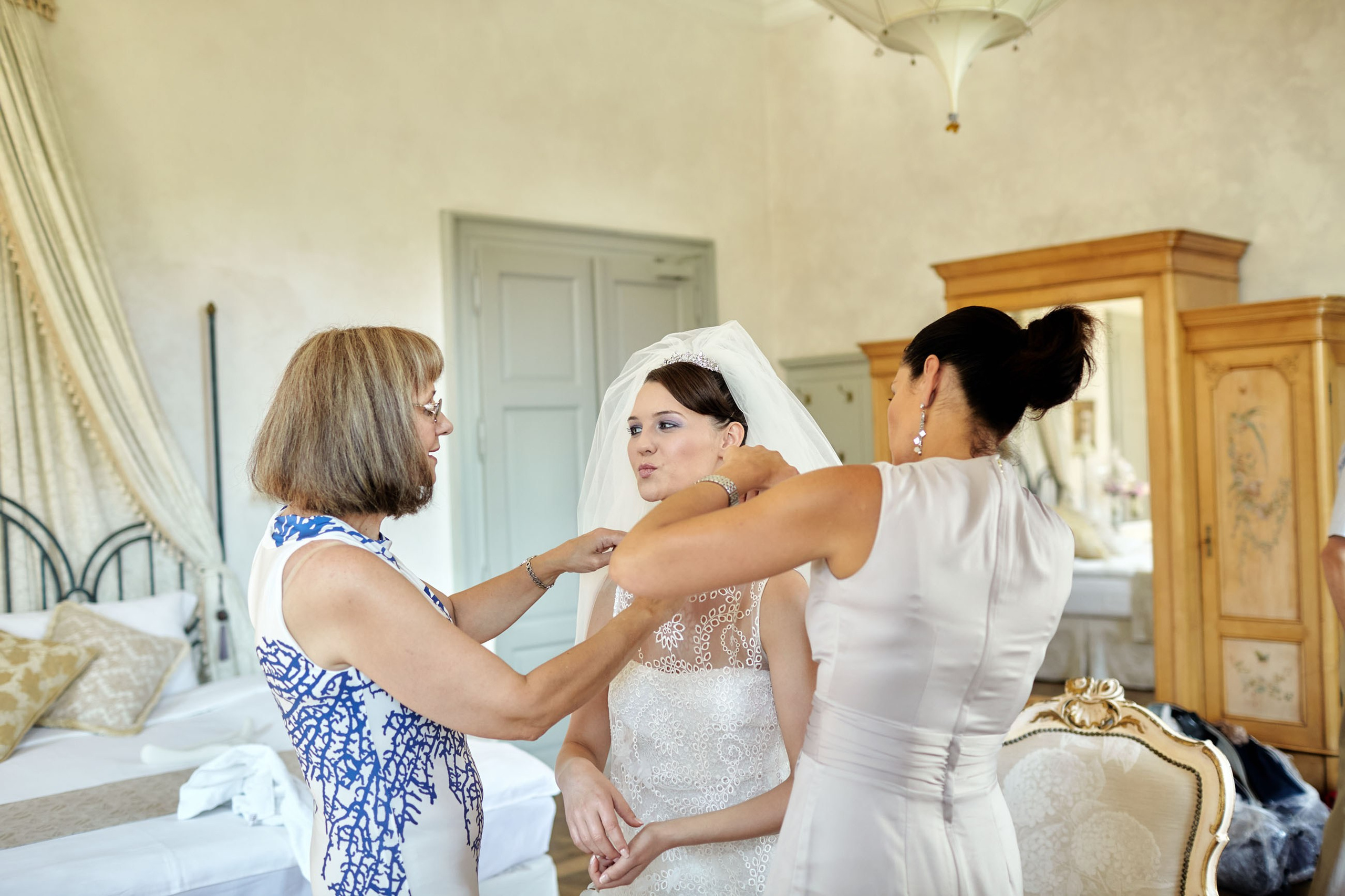 Bride mimicks kiss for her mother during veil adjustment in historic chateau.