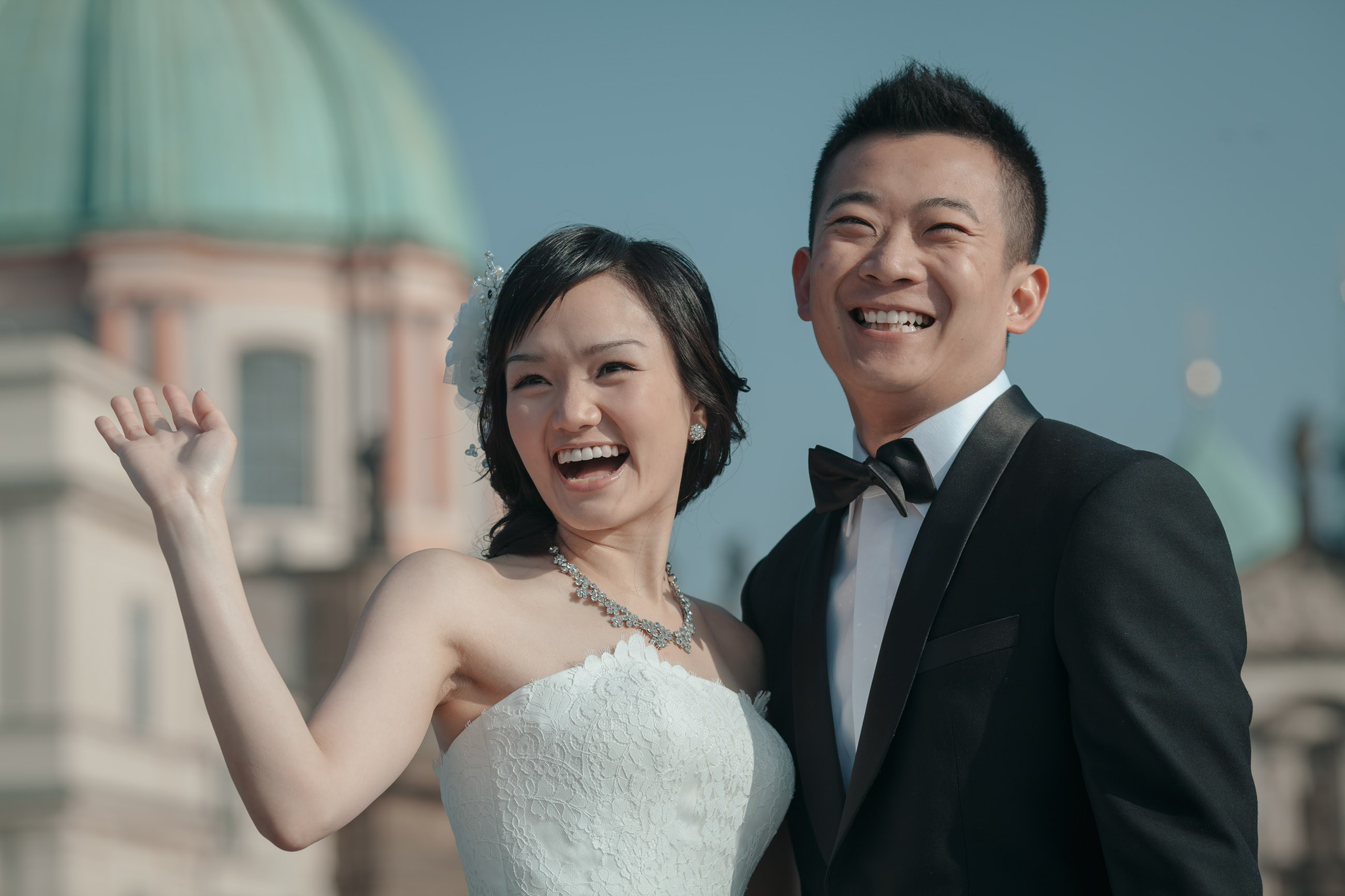 An elegant couple from Hong Kong dressed in their stylish wedding attire wave to onlookers as they pose for a photo during their Prague pre-wedding atop the Charles Bridge.