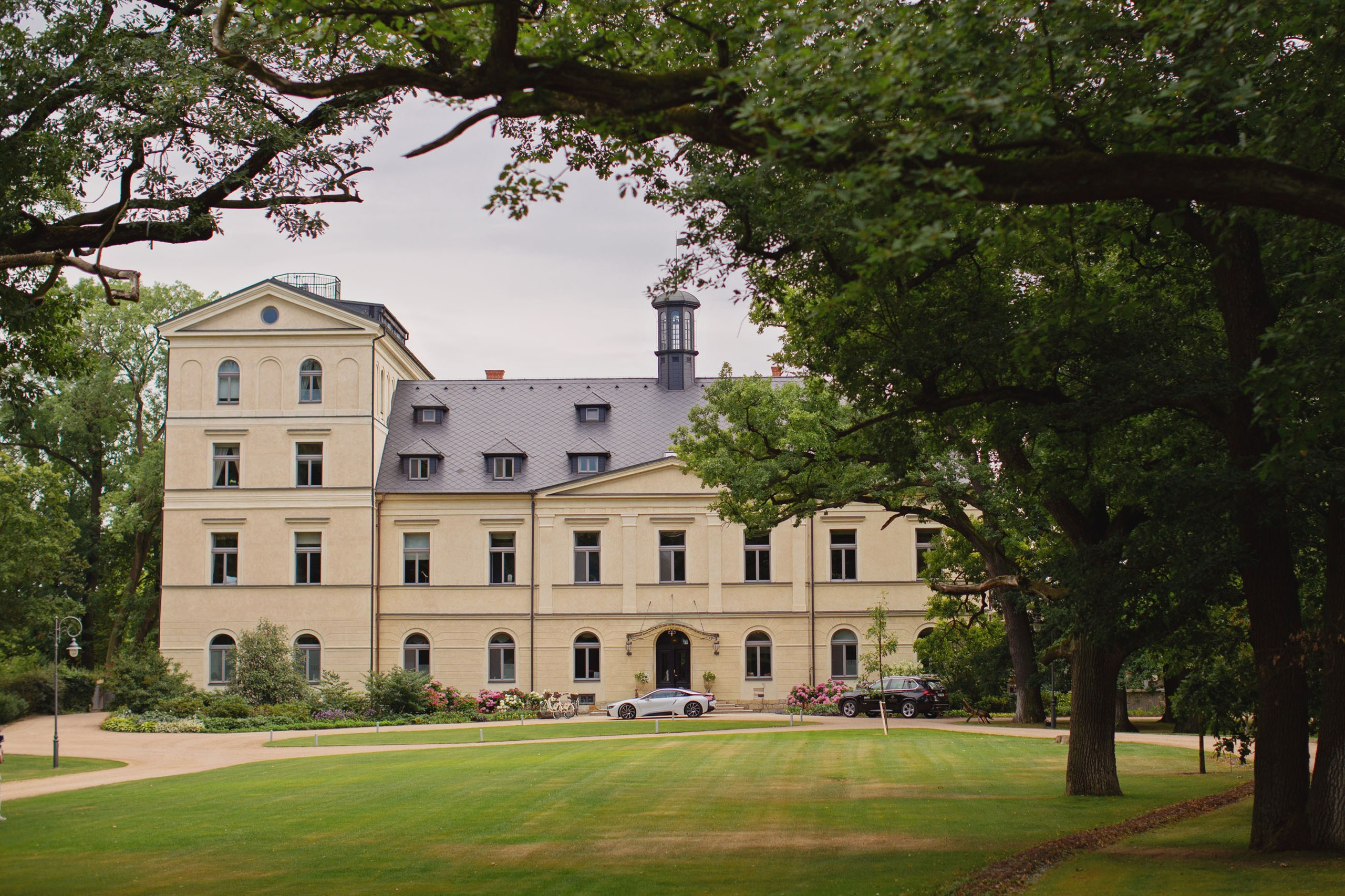 Exterior view of Chateau Mcely's historic facade with luxury cars parked on its beautifully landscaped grounds.