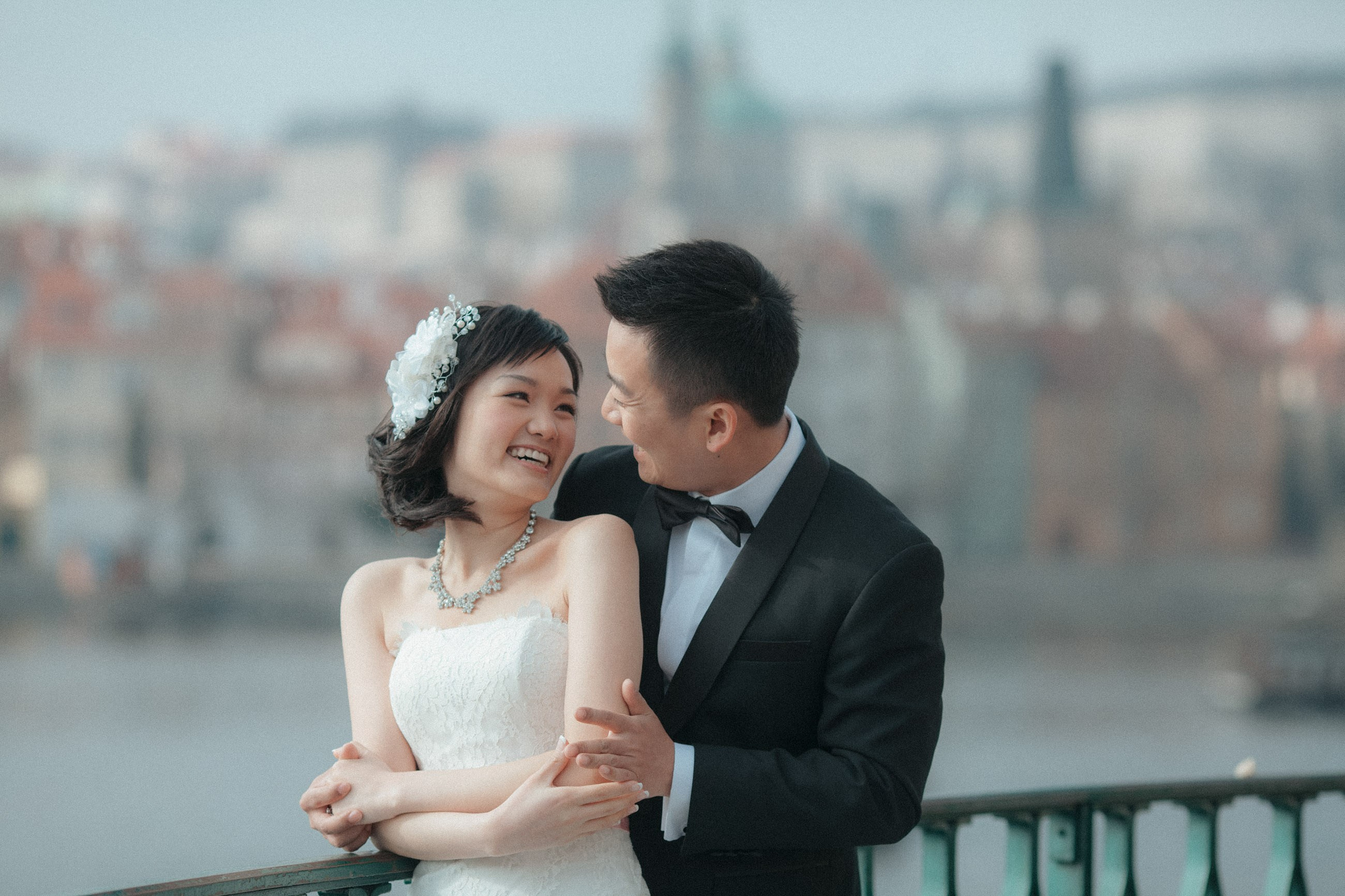 A tuxedo wearing Hong Kong groom and the beautiful bride laugh as they snuggle up for a wedding portrait near the riverside in Prague.
