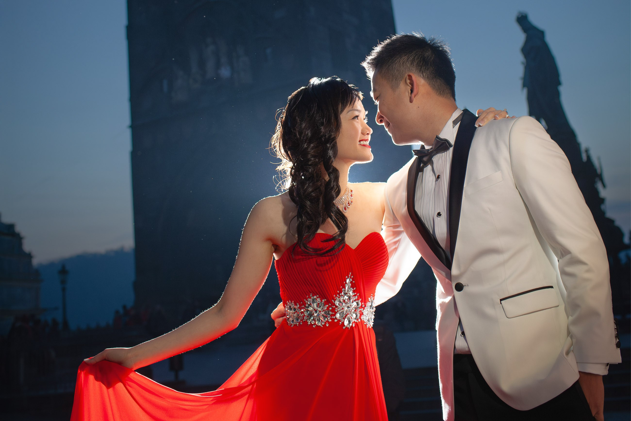 As the powder tower above the Charles Bridge towers above, a stylishly dressed Hong Kong woman in a red dress is caressed by her white tuxedo wearing fiance in Prague.