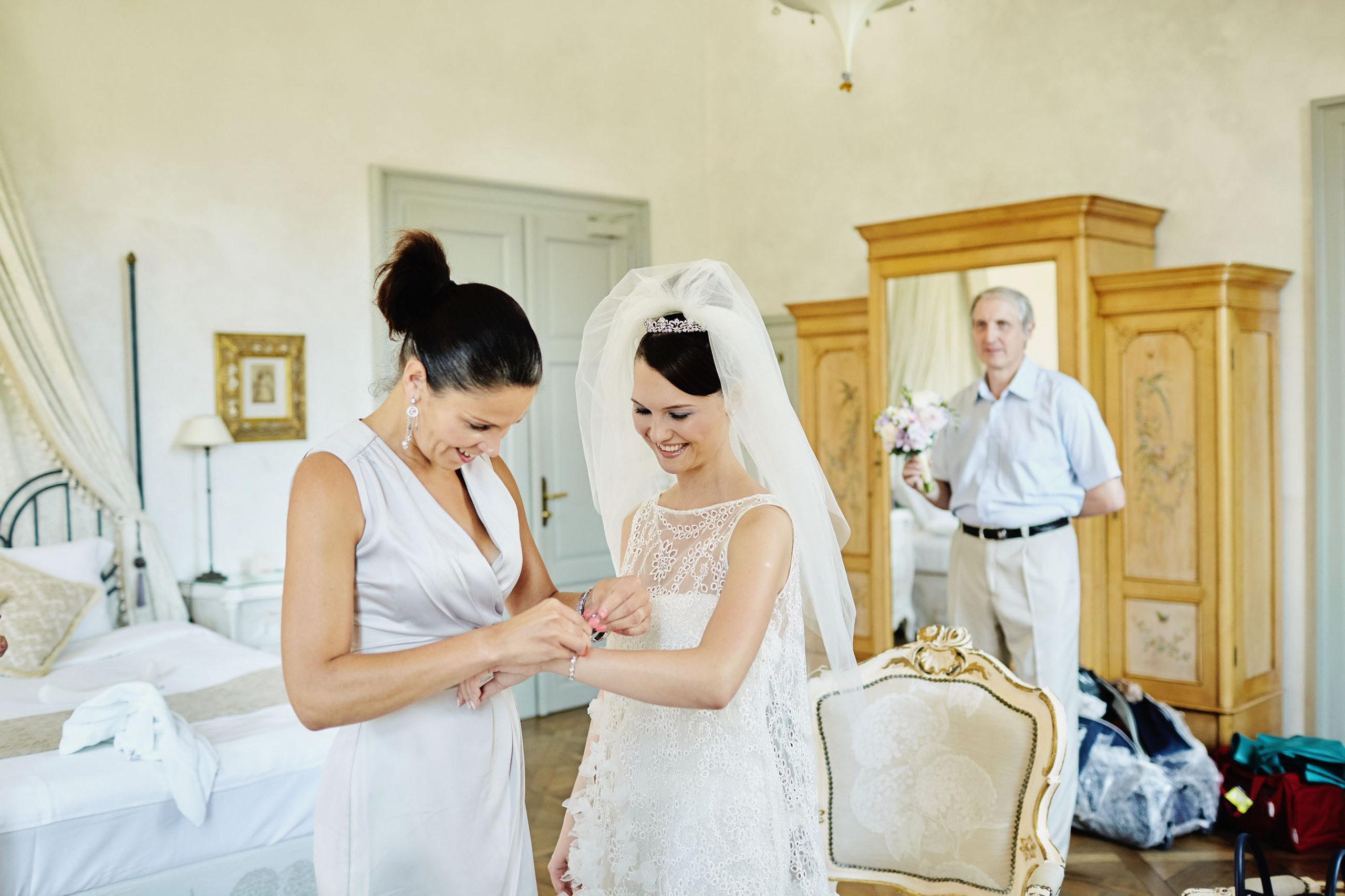 Makeup artist adjusting bride's bracelet as father with bouquet observes at historic chateau.