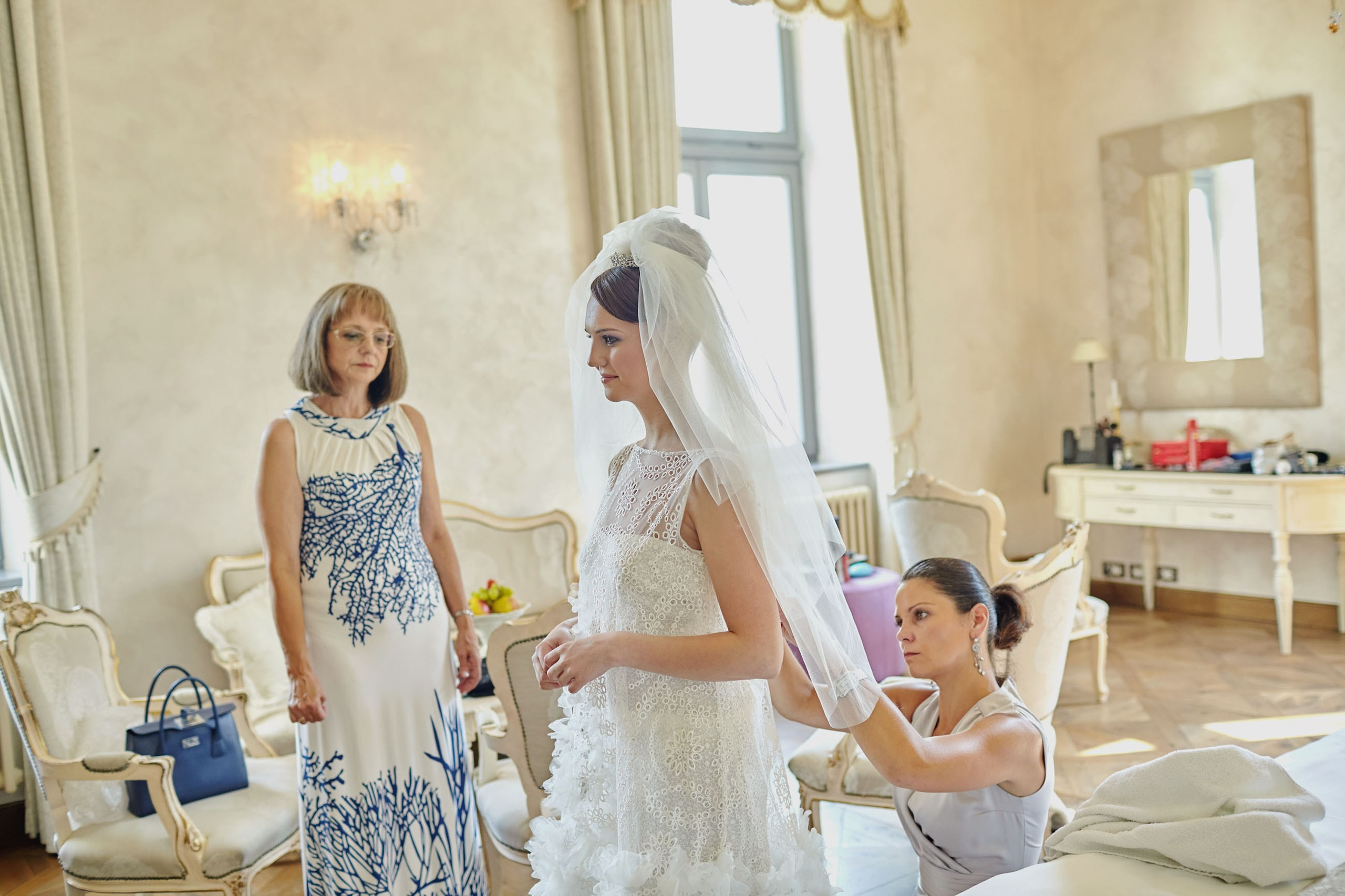 Makeup artist adjusting bride's dress under her mother's watch in Chateau Mcely's bridal suite.