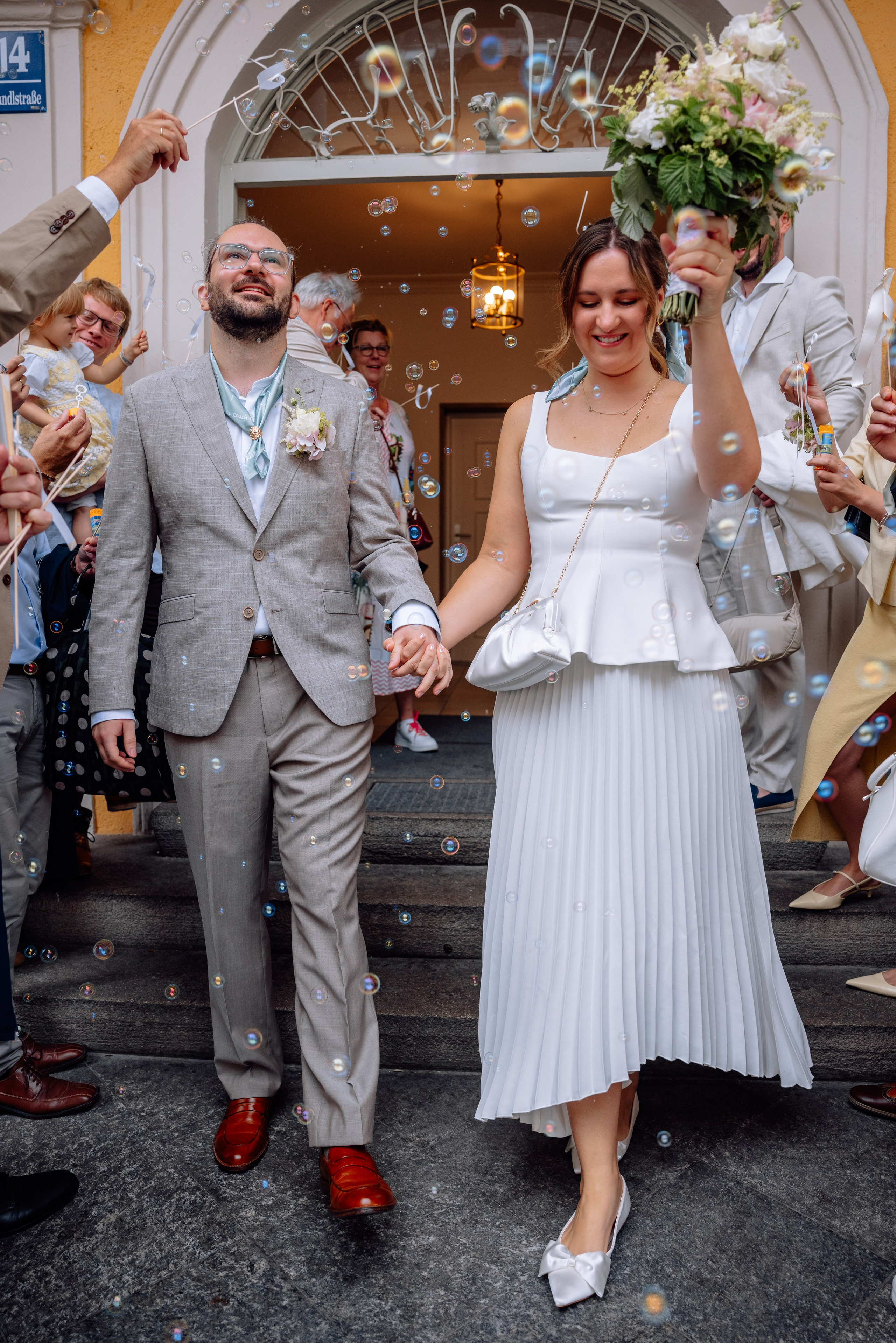 Braut trägt großen Blumenstrauß – Paar verlässt Standesamt Mandlstraße 14, geht im Park Hand in Hand.