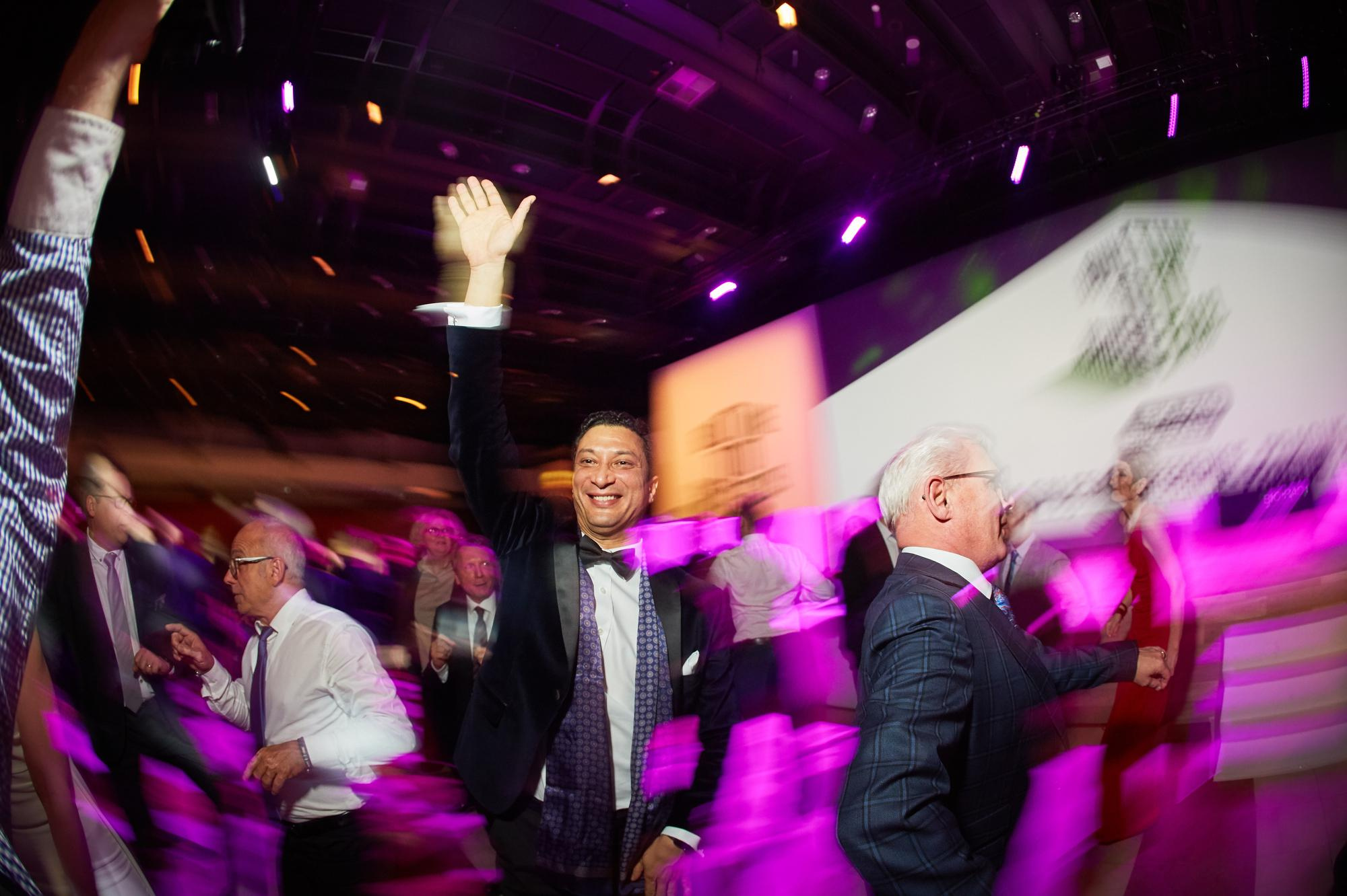 A tuxedo-wearing executive waves to onlookers under the colorful lighting as a band performs on stage.