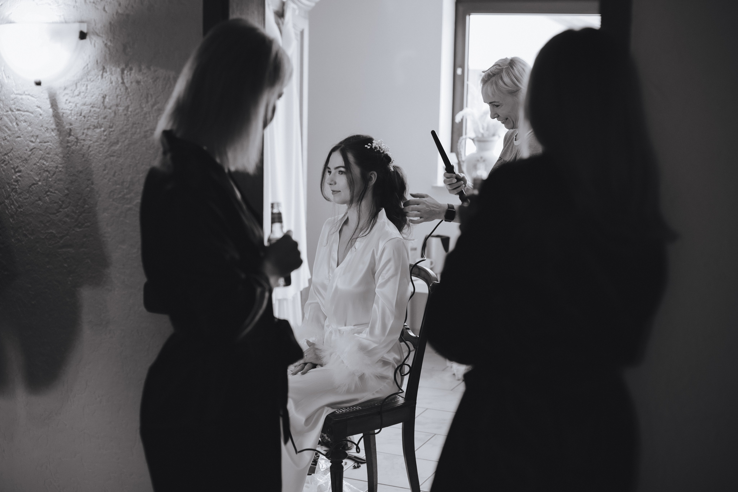 Die Brautjungfern schauen neugierig durch die Tür, während der Braut die Frisur gemacht wird – sie lächelt aufgeregt und voller Vorfreude.