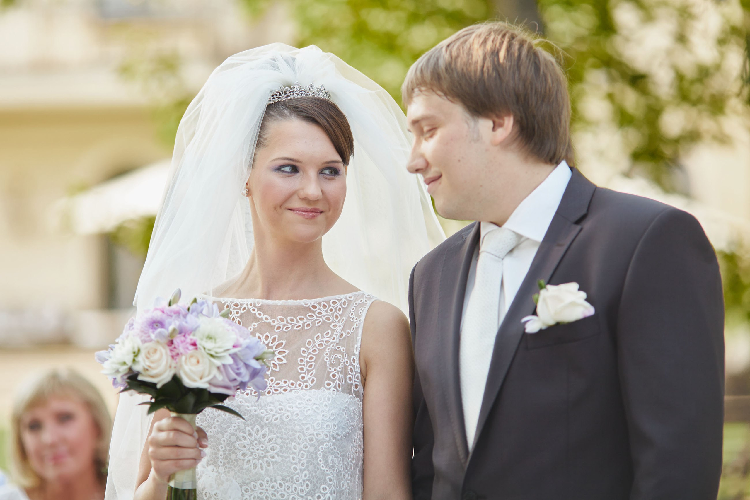 The bride, holding her bouquet, looks towards the groom and smiles during their outdoor garden wedding on the grounds of a historic chataeu in Czechia.