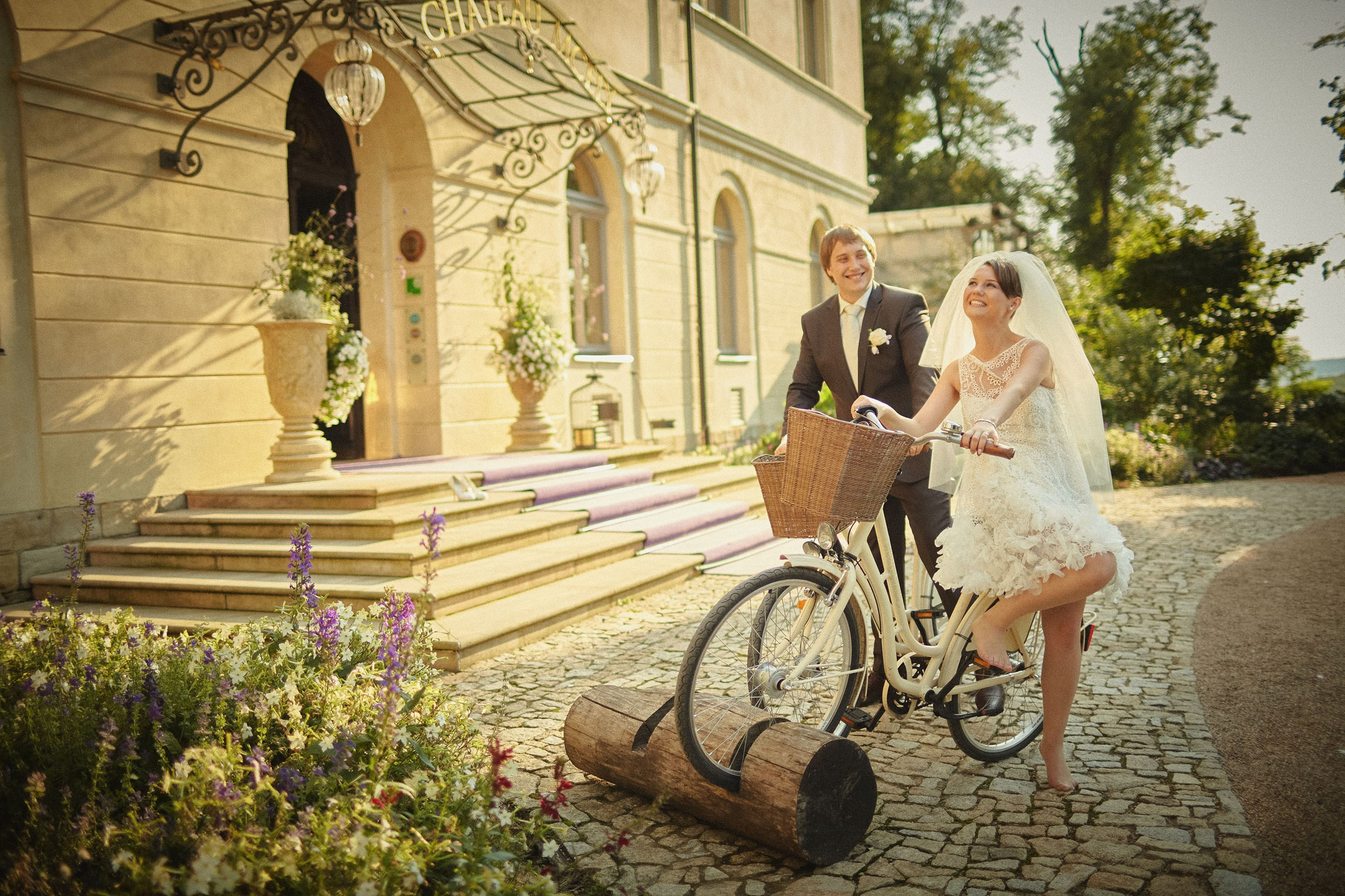 Young newlyweds atop bikes near the grand entrance of Chateau Mcely laugh out loud as they share a funny moment on their wedding day.
