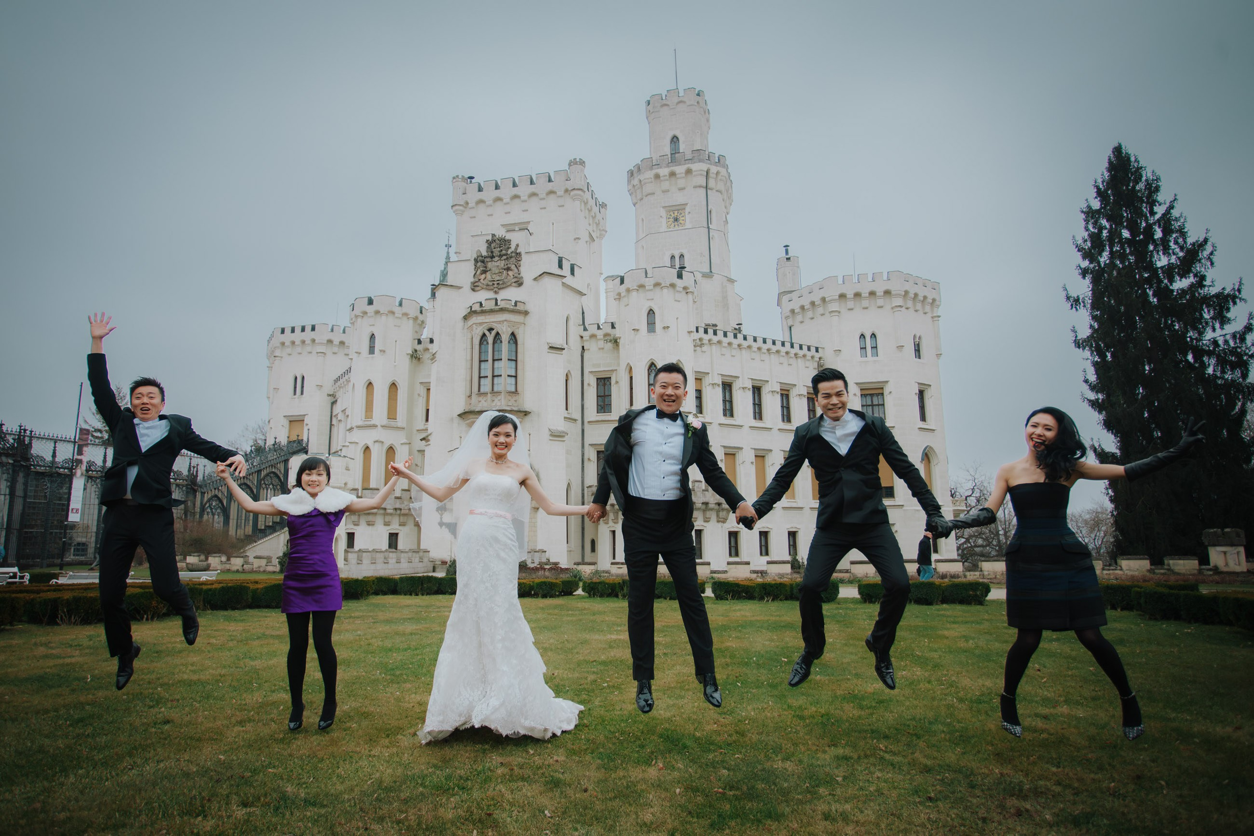 A smiling bride & groom from Hong Kong are joined by their friends as they hold hands and jump high into the air on the grounds of the State Chateau of Hluboka during their wedding day.