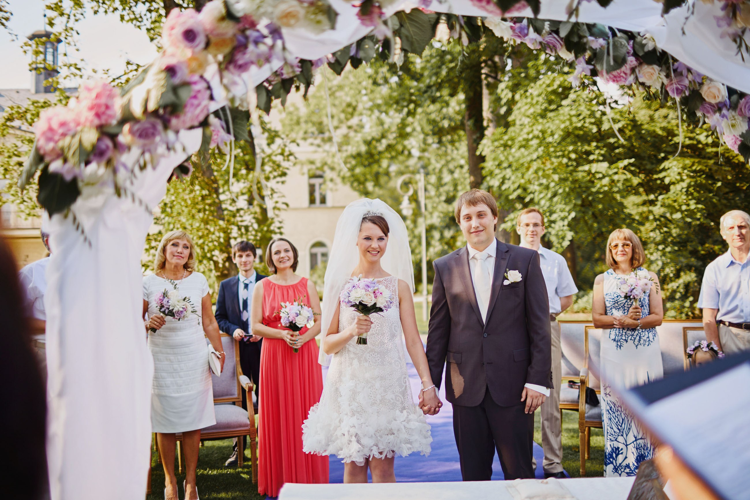 Smiling bride and groom under floral arch with guests at chateau wedding.