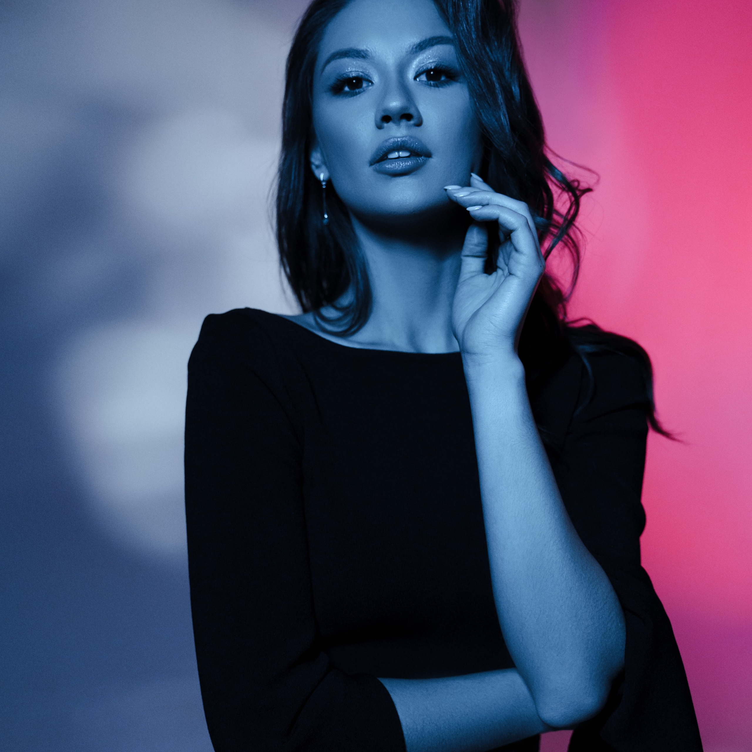 Studio portrait of a woman in a black dress, illuminated by moody blue and pink lighting. High-fashion editorial photoshoot, beauty photography.