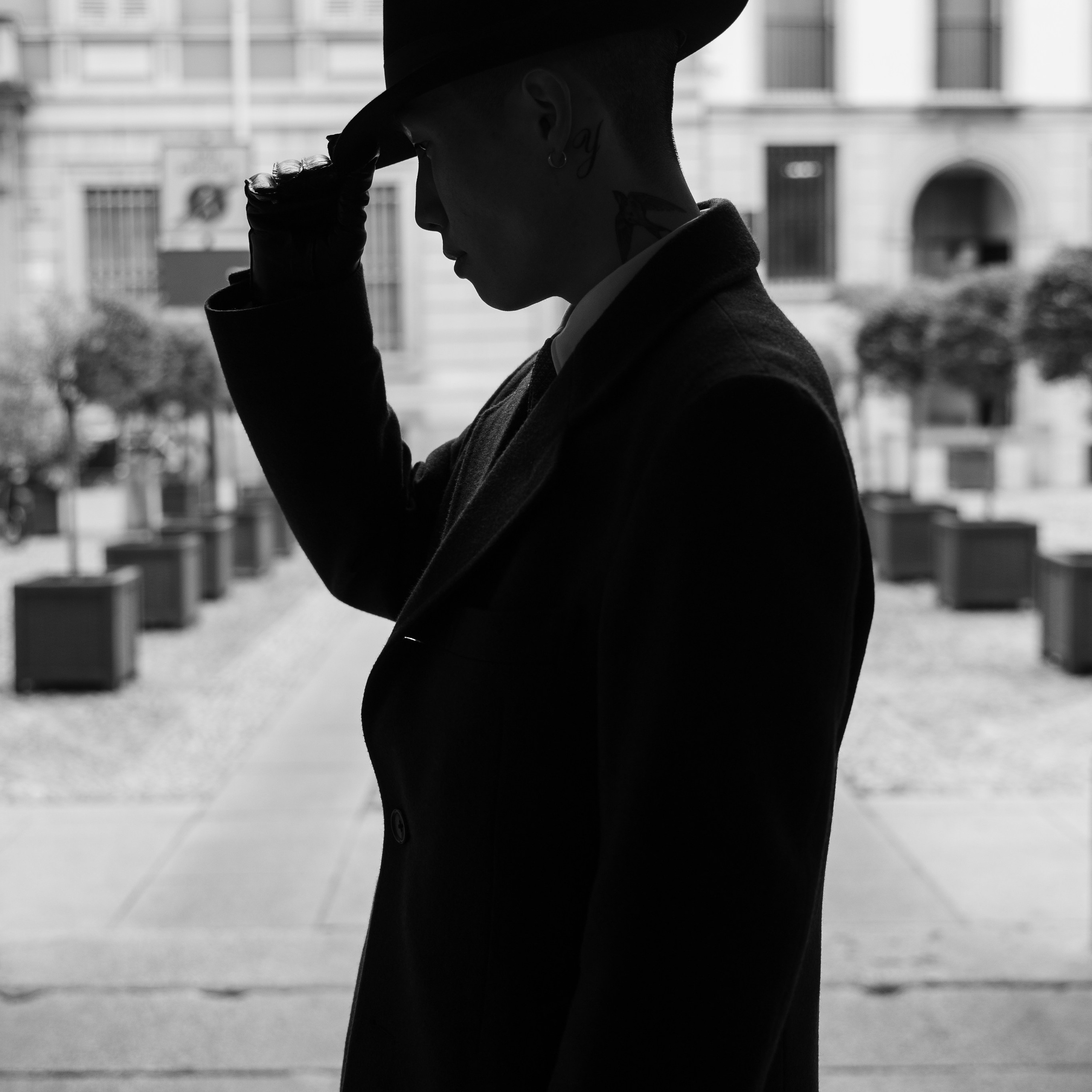 Dramatic black-and-white silhouette of a man in a fedora hat, adjusting his brim in front of a classical Milanese building