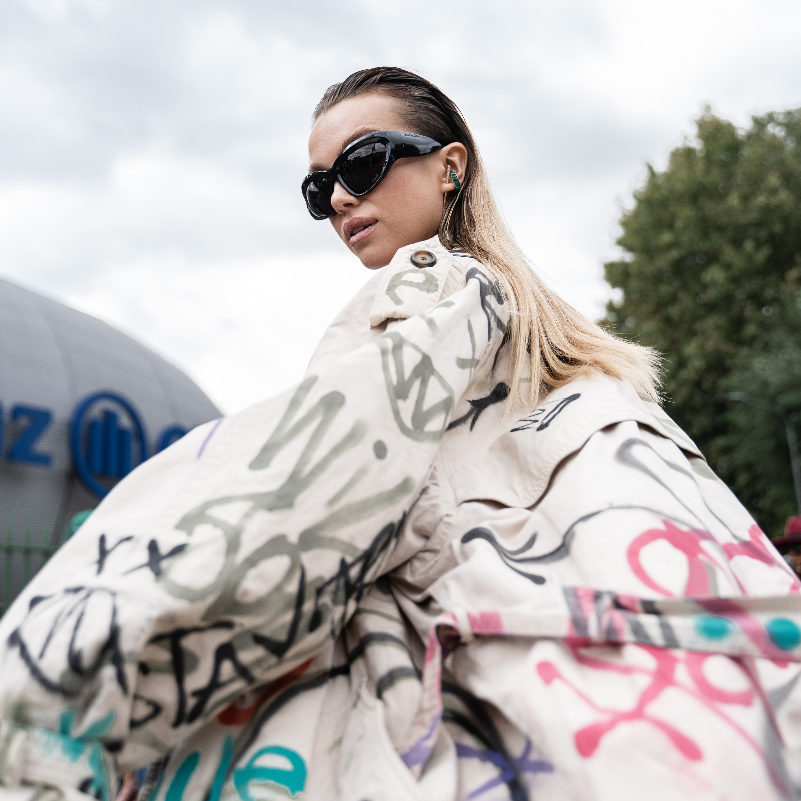 Fashion-forward woman in an oversized graffiti trench coat and sunglasses seen at Milan Fashion Week