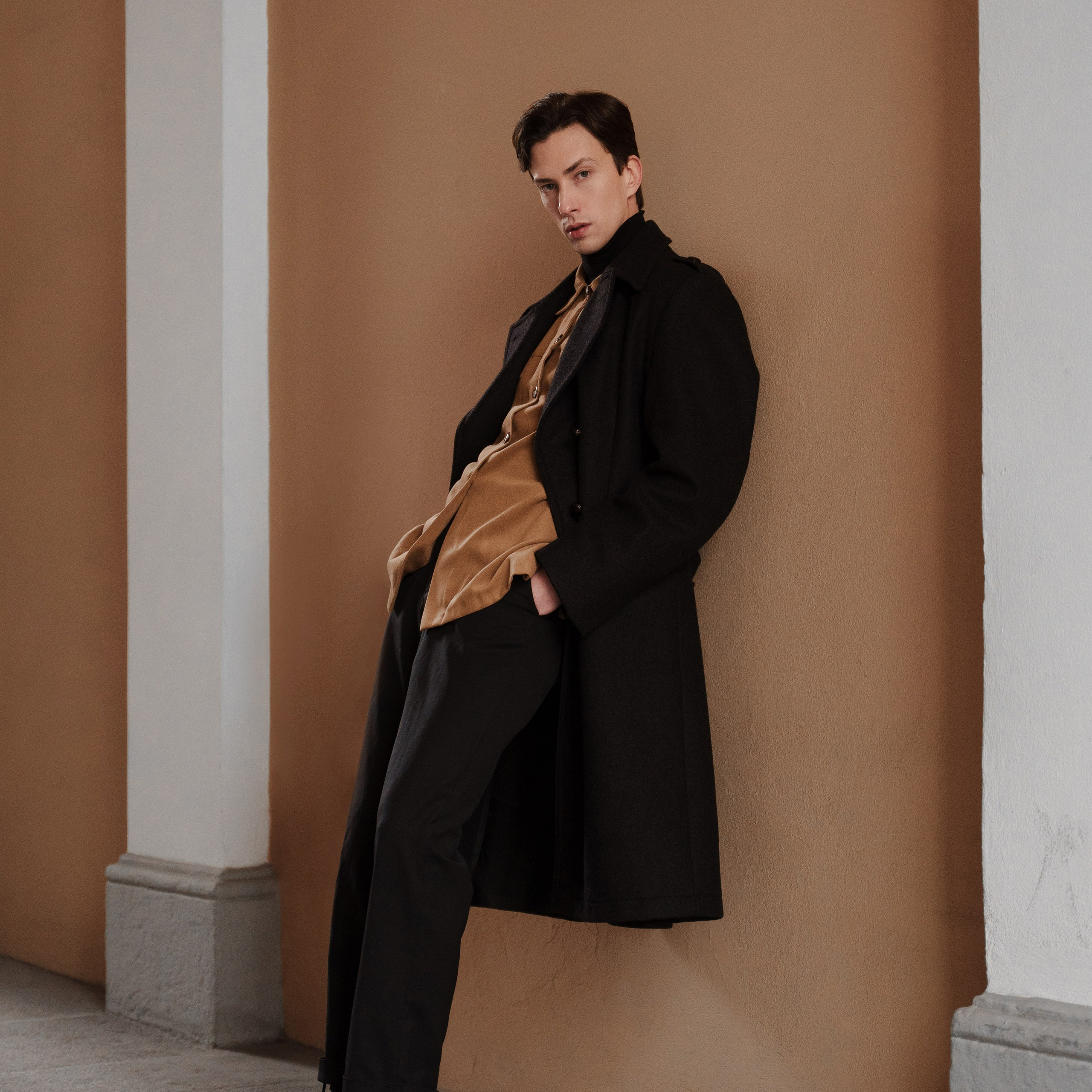 Elegant young man leaning against a peach-colored wall, dressed in a sophisticated dark coat over a camel jacket and black turtleneck, complemented by stylish black trousers and chunky lace-up boots, exuding a sense of modern dandyism.