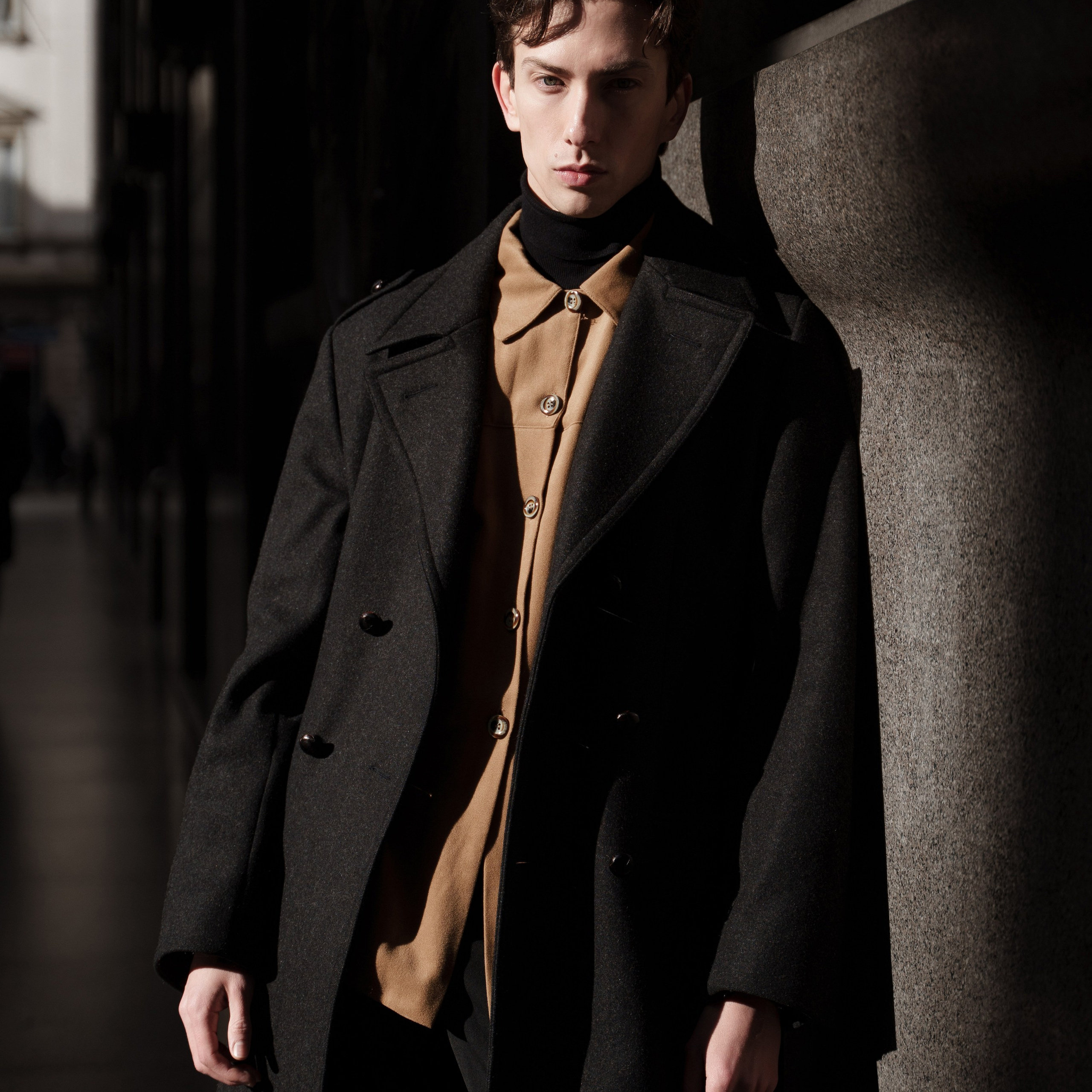 Portrait of a young man in elegant autumn fashion, wearing a camel turtleneck and a dark overcoat, partially illuminated by natural light on a city street