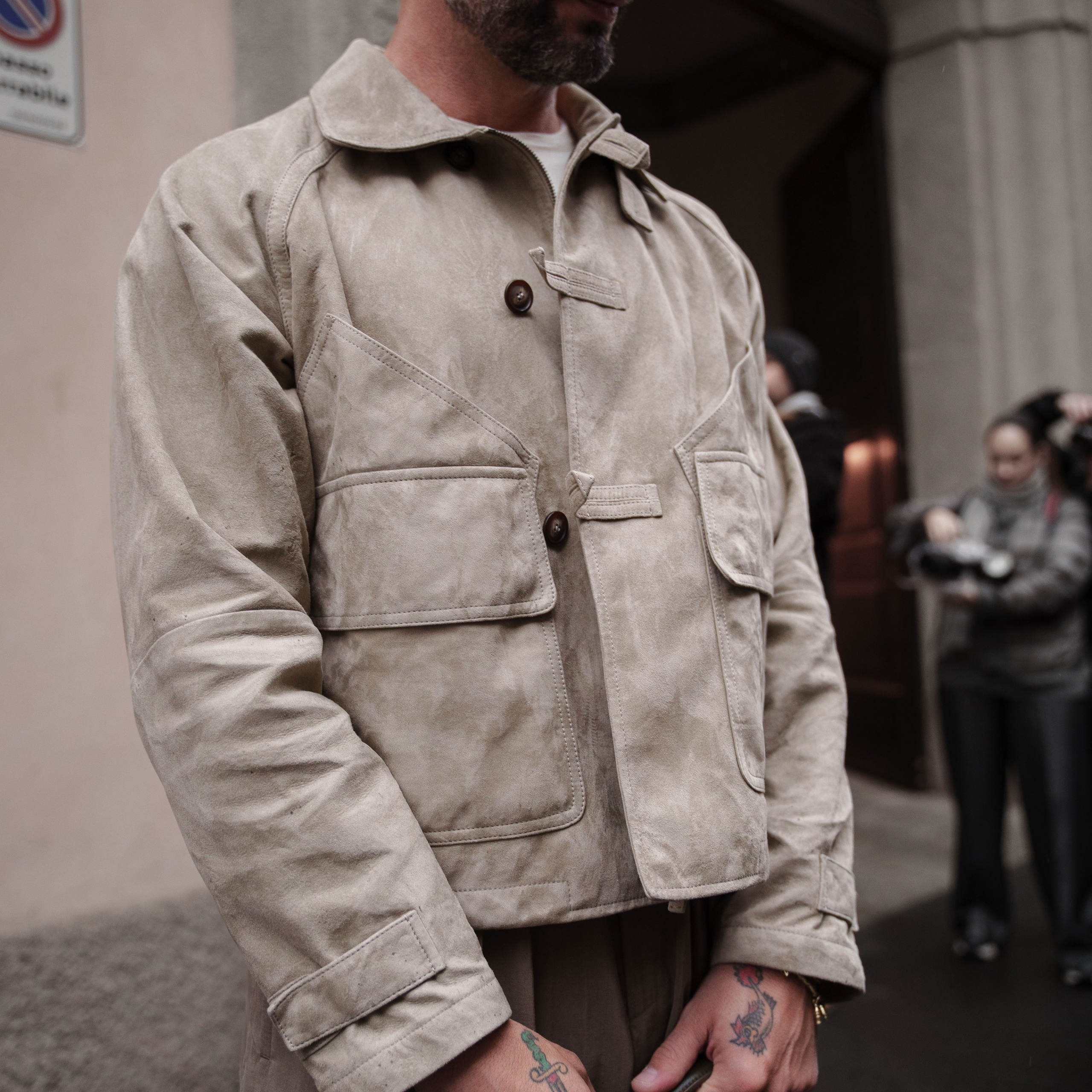 Milan Fashion Week photographer captures a man in a beige suede jacket with tattoos and sunglasses on the streets of Milan