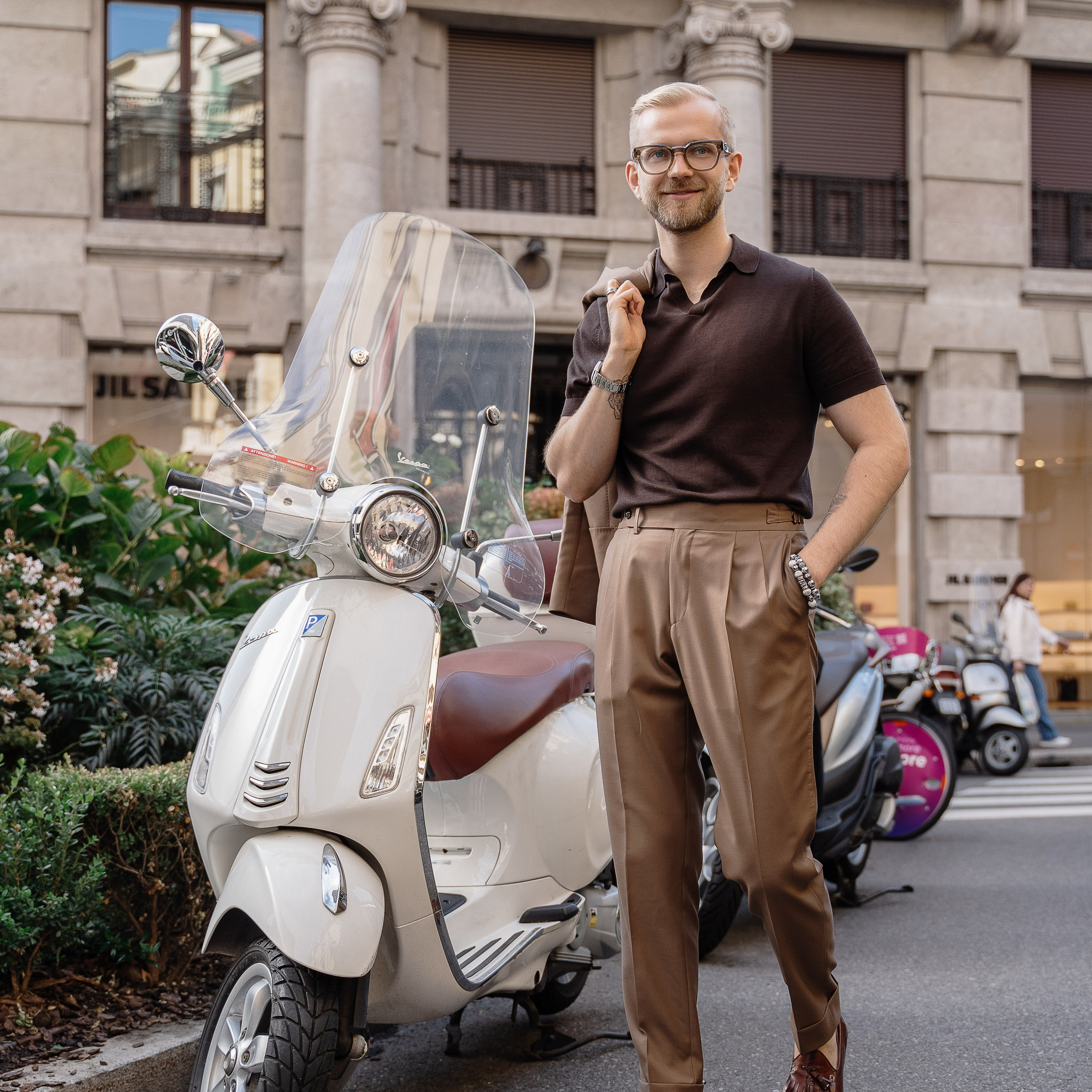 Chic man in a brown polo and tailored trousers standing next to a classic white Vespa in a Milan street