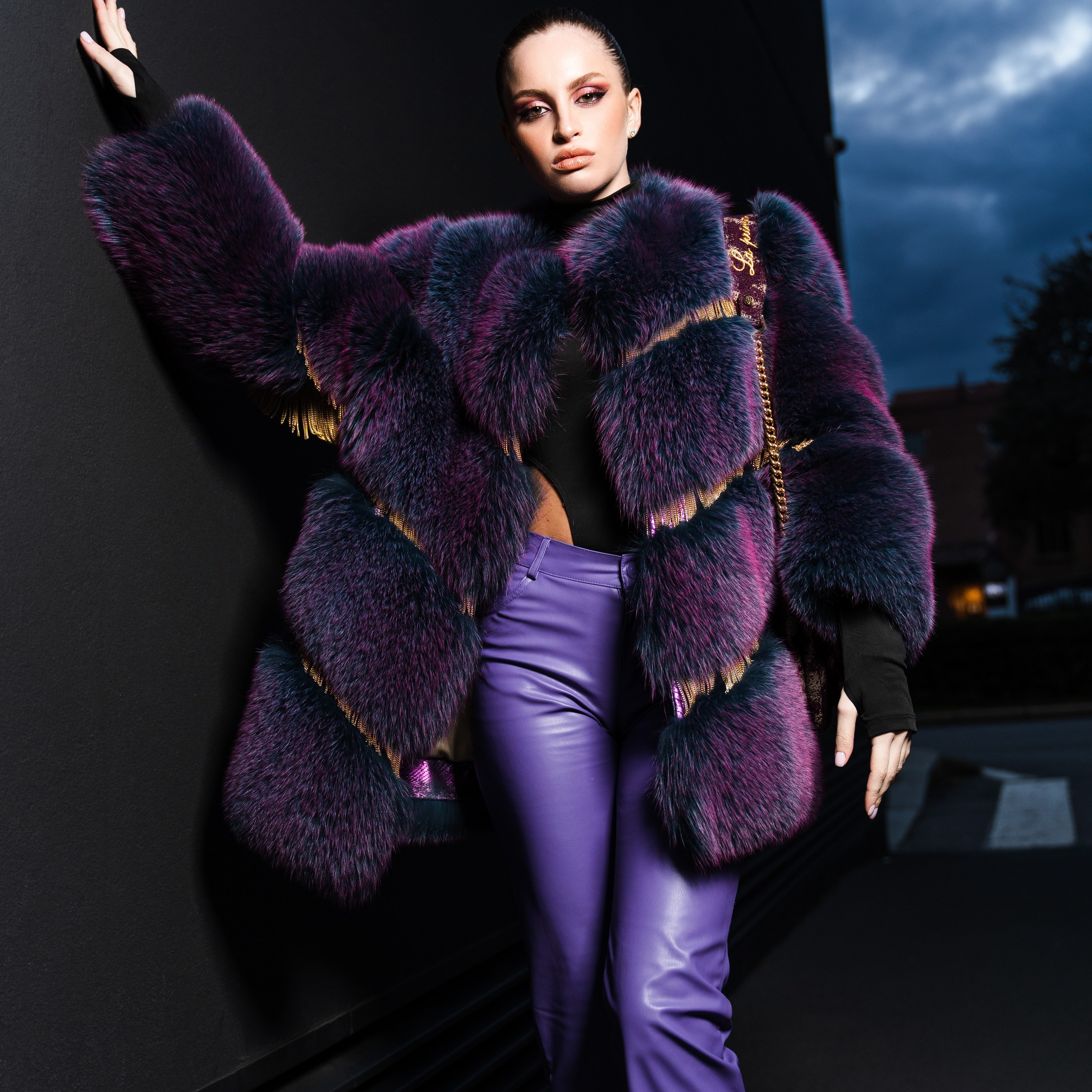 High-fashion portrait of a woman in a bold purple fur coat and leather pants, posing against a dark urban backdrop at dusk