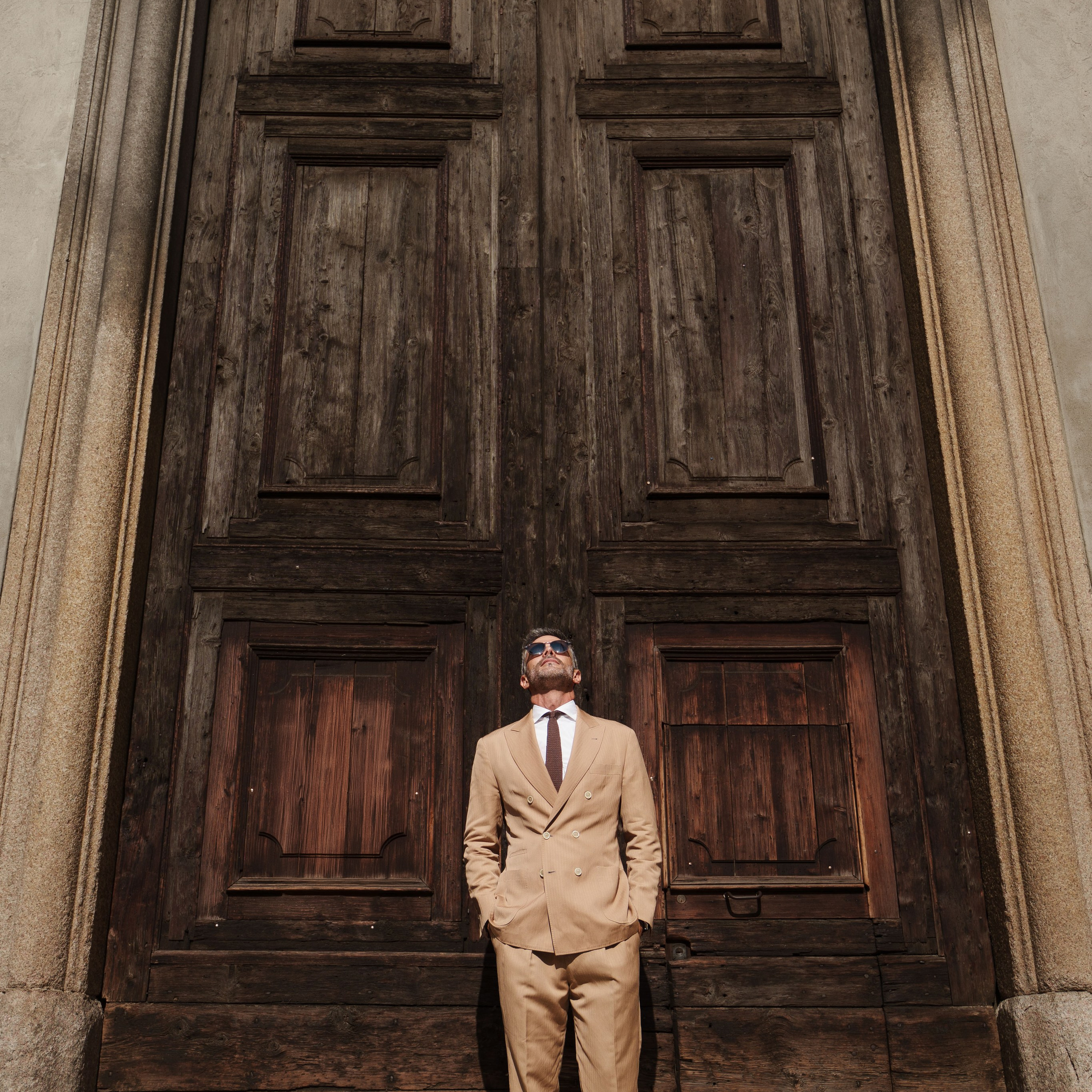 Fashion editorial shot of a man in a beige suit, posing in front of a grand historic wooden door in Milan. Street photography, architectural portrait