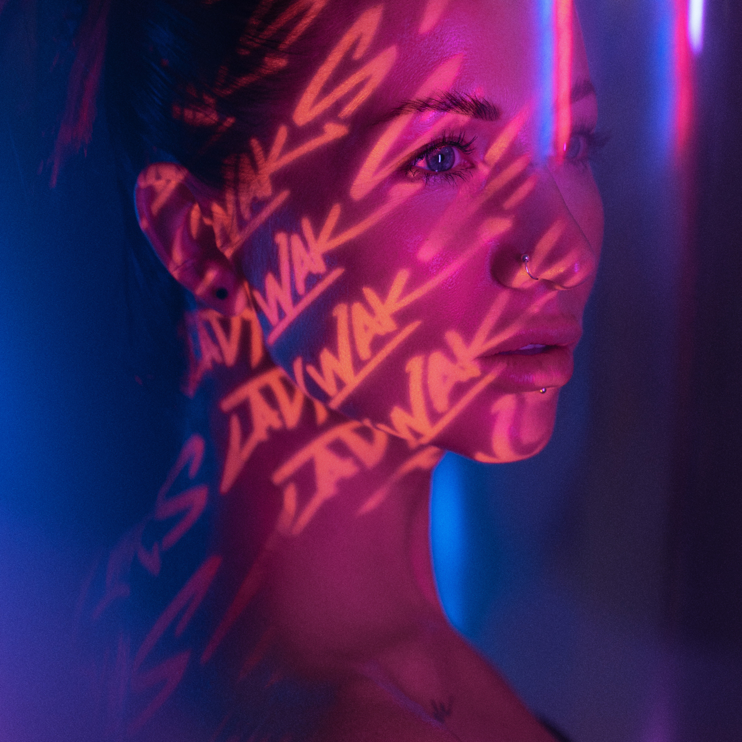 Artistic portrait of a woman with vivid neon light patterns cast across her face, highlighting her piercings and tattoos