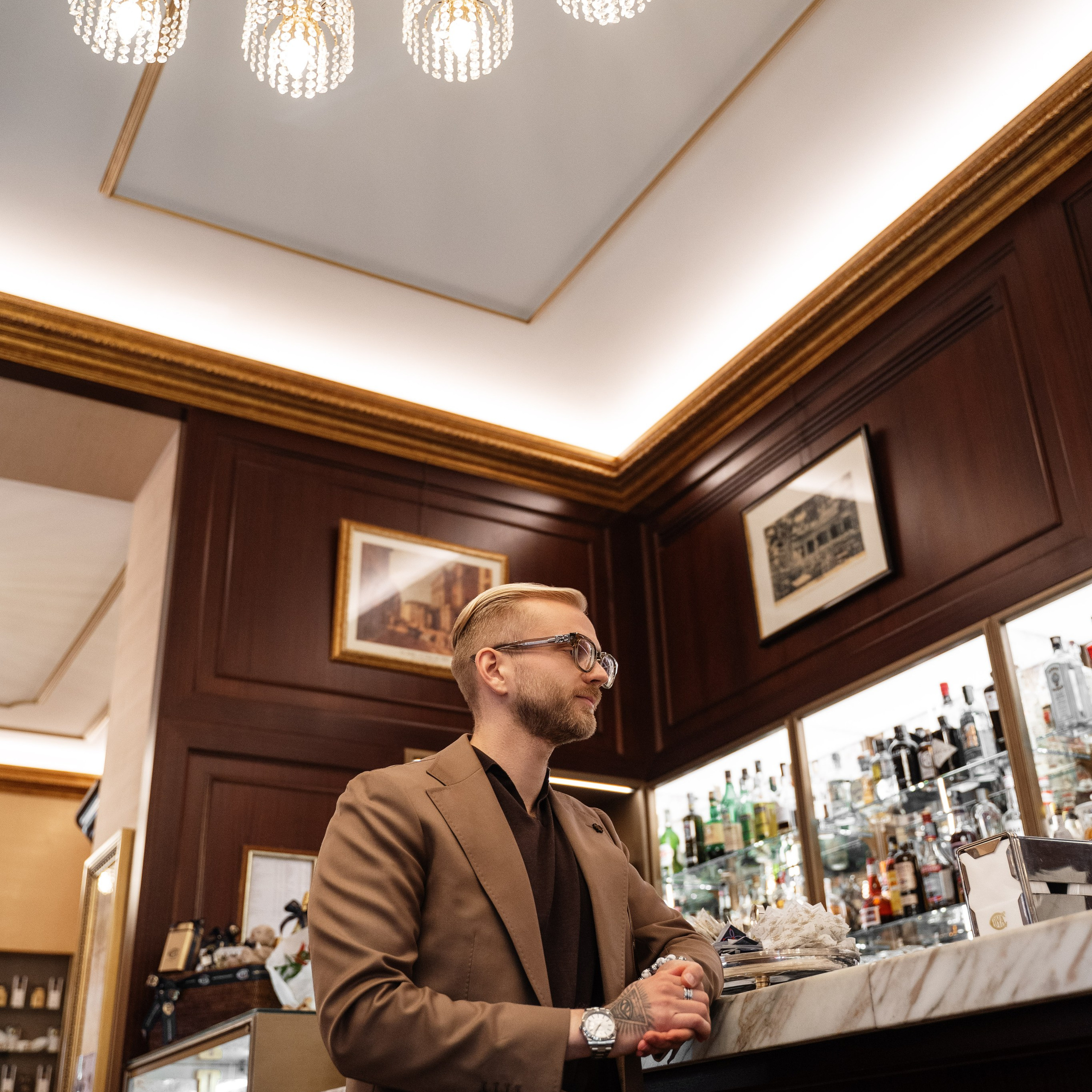 Lifestyle portrait of a sophisticated man in a brown suit, standing under a grand chandelier in a Milanese café. Fashion photography, luxury editorial