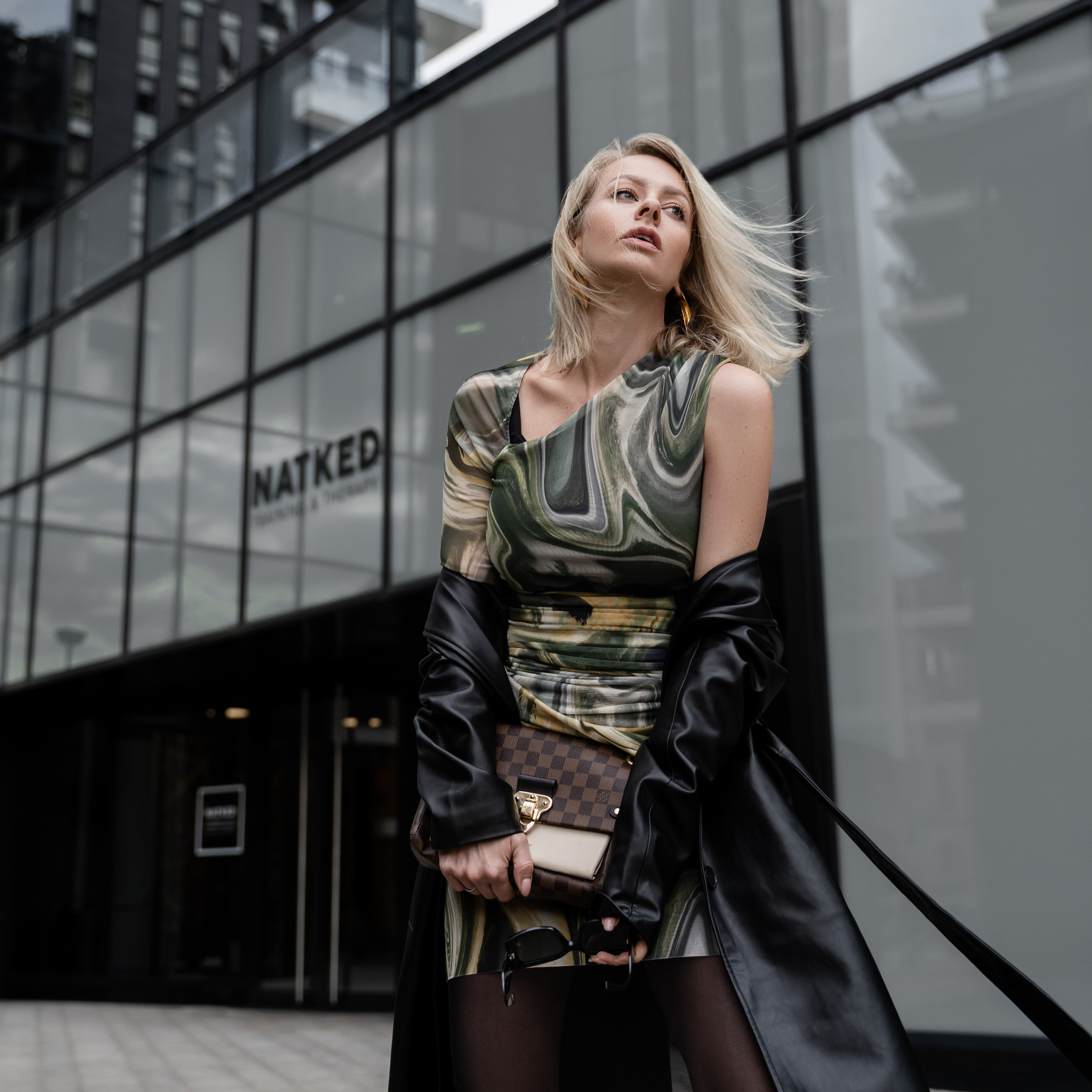 Fashion-forward woman in a patterned dress and black coat posing in front of a glass building