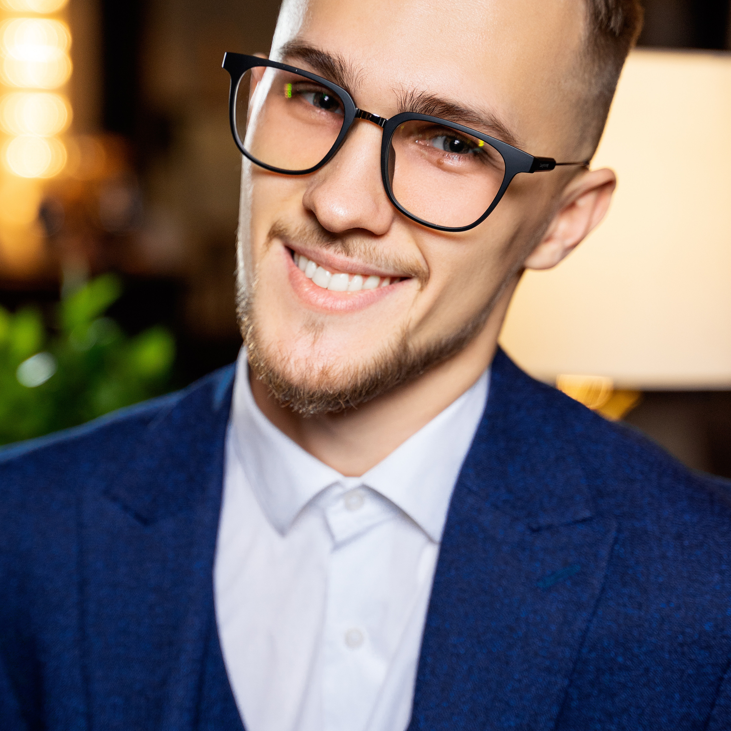 Smiling young man with a neat beard wearing glasses and a blue suit in a bright interior