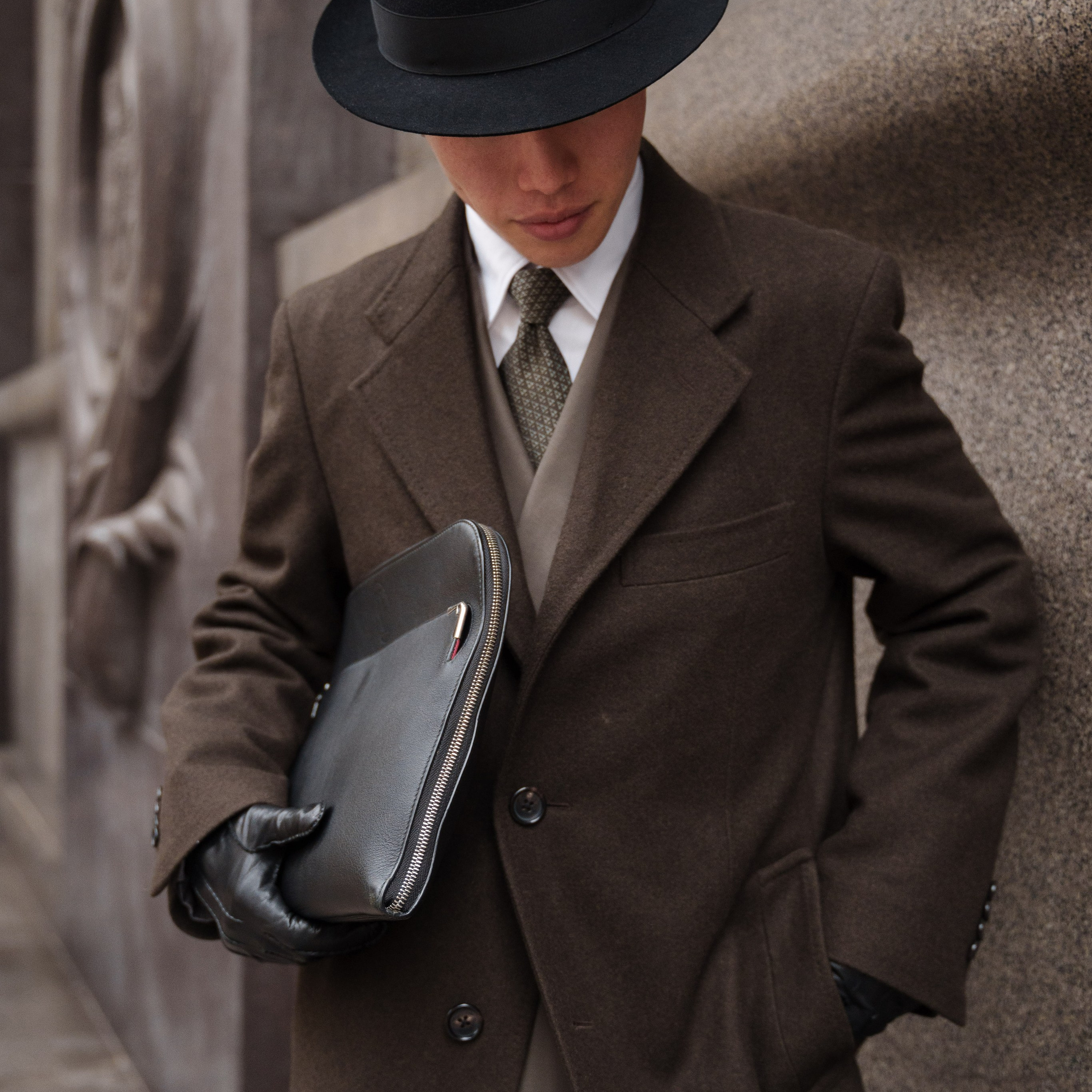 Sophisticated man in a brown wool coat and black fedora hat, standing near a textured stone wall in Milan