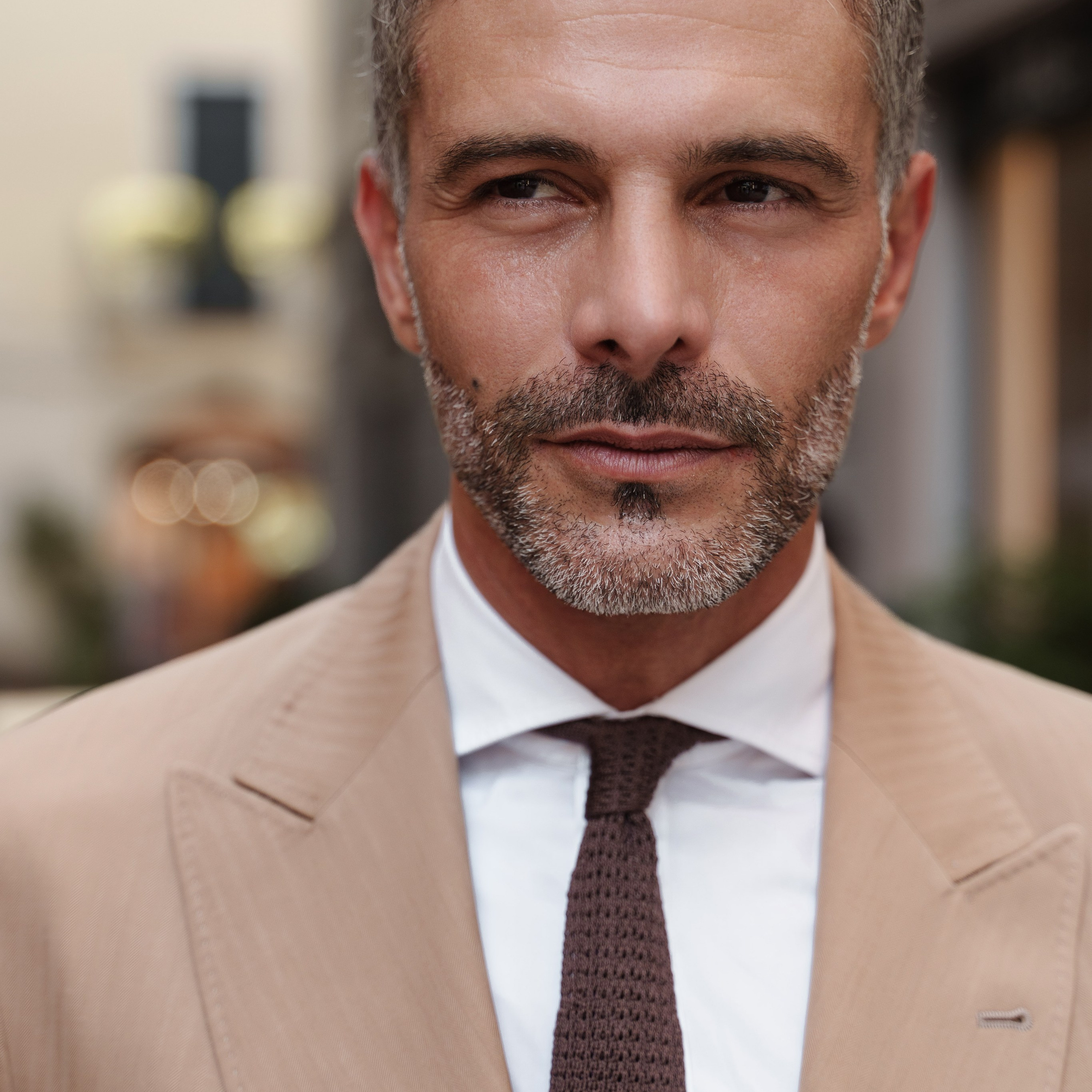 Close-up portrait of a confident middle-aged man with salt-and-pepper hair, wearing a beige suit and brown tie, captured in a Milan street